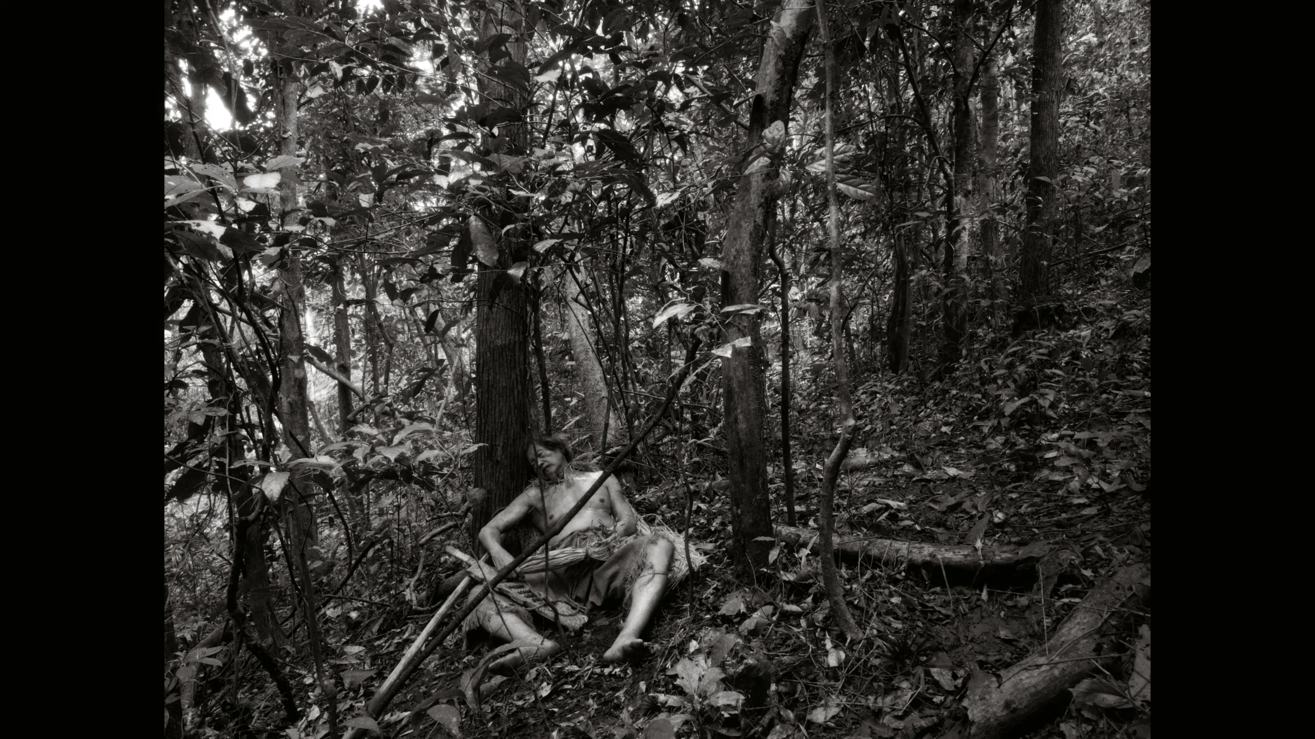 Shirtless person seated on the forest floor among tangled brush and tree trunks in a black-and-white image.