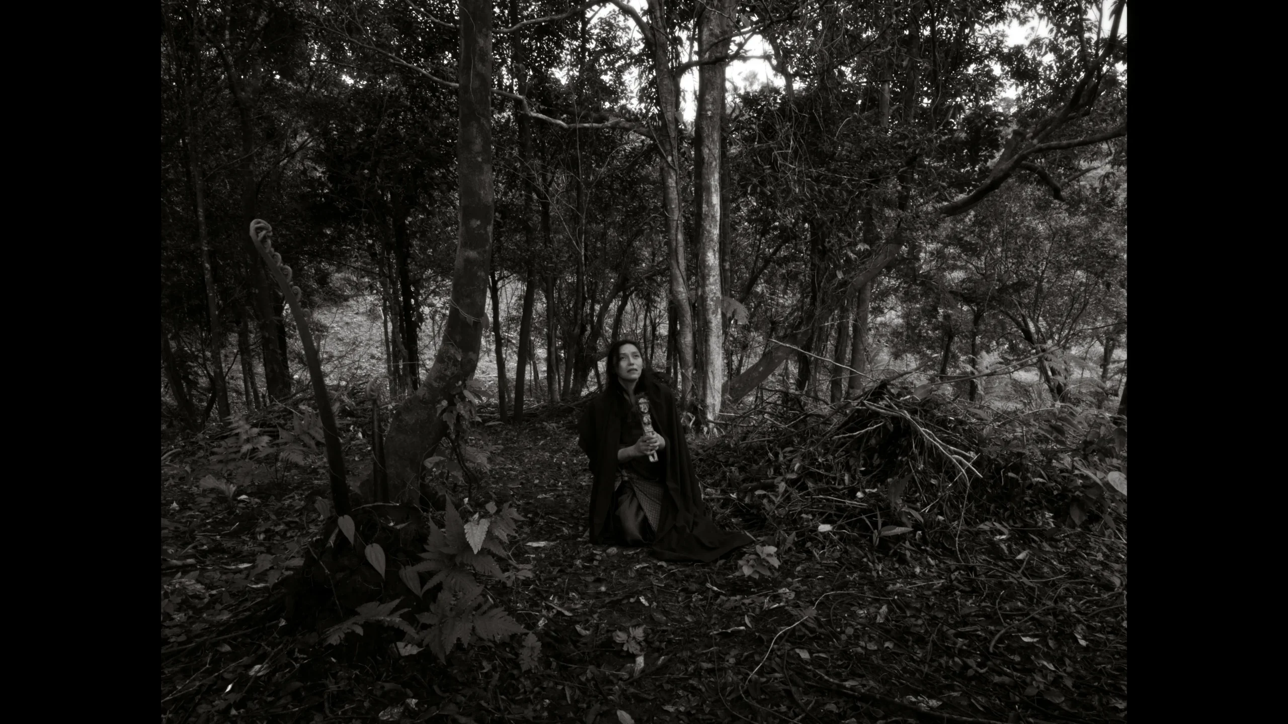 Woman kneels in a dense forest undergrowth, wearing dark clothing and holding a carved staff or object, amid bare tree trunks.