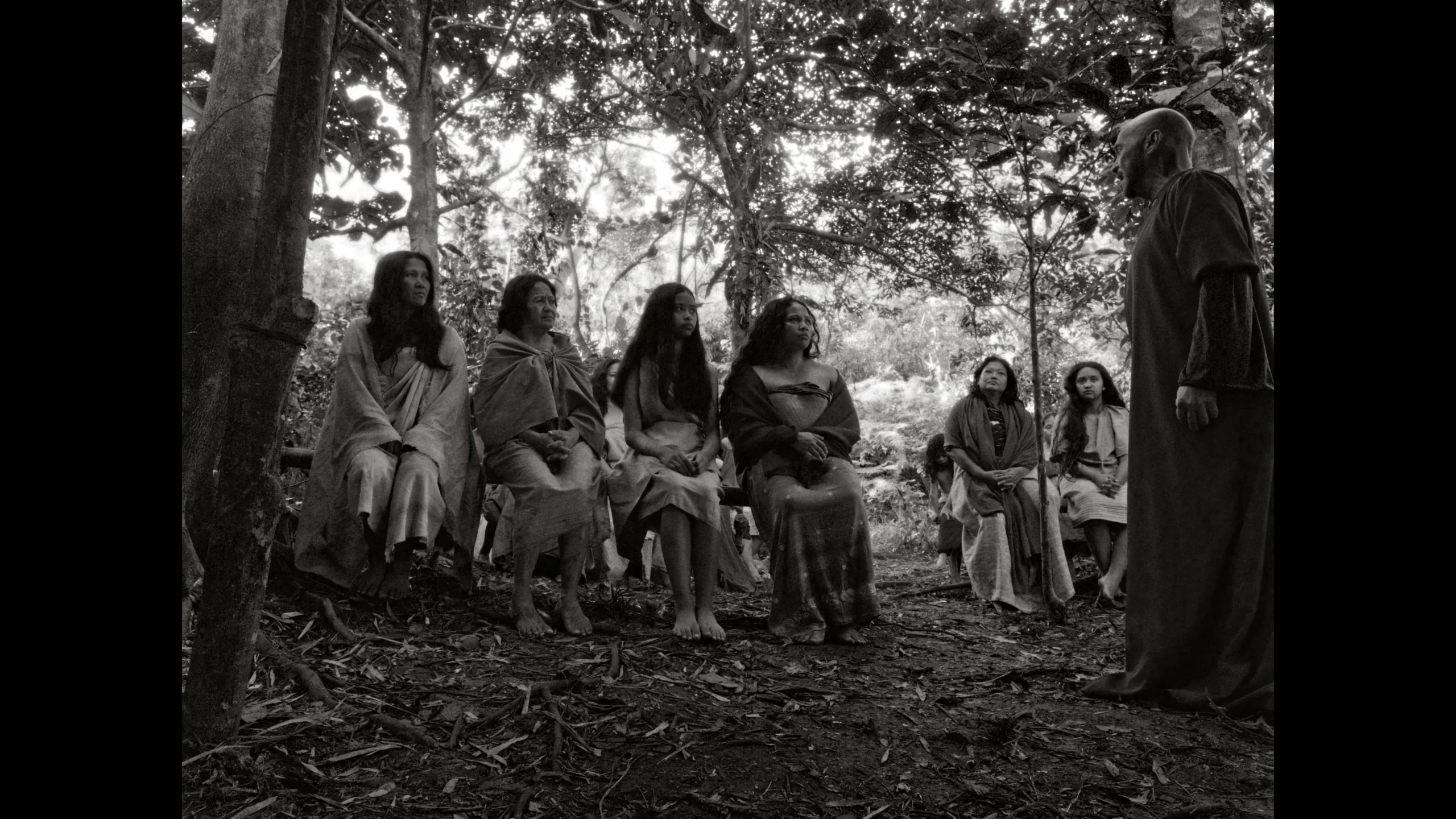 Group of people seated on a log in a forest, listening to a standing man in robes (black-and-white photo).