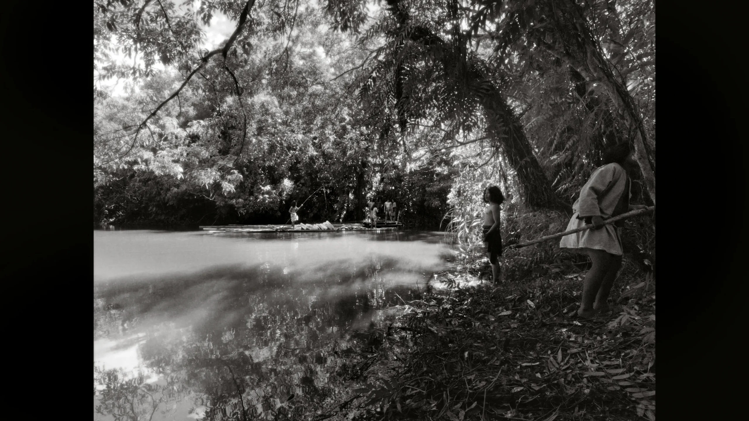 Children by a riverside fishing with sticks while a canoe drifts on calm water, surrounded by dense trees.