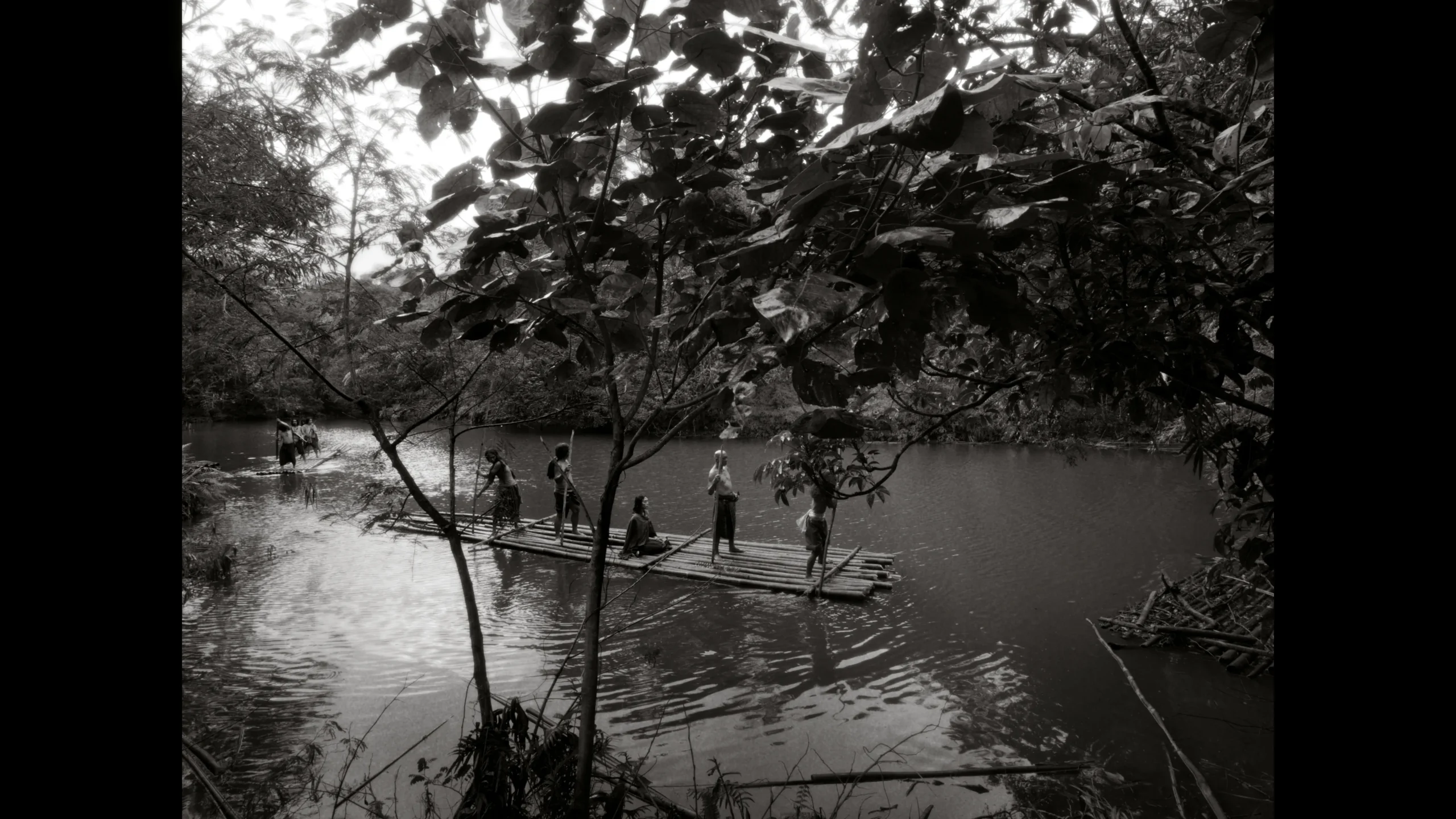 Children stand and sit on a long bamboo raft drifting on a calm river, surrounded by dense trees and foliage.