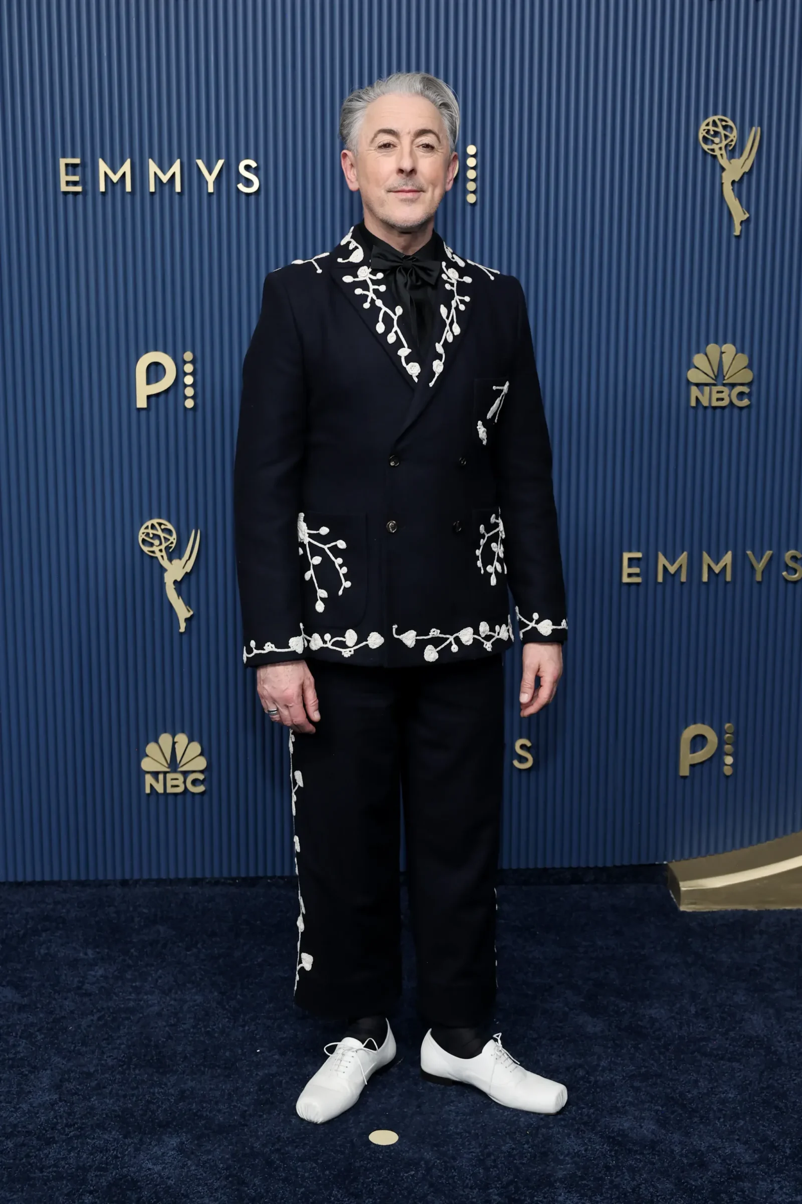 Man in a dark suit with white floral embroidery poses on a blue Emmy backdrop with NBC logos nearby.