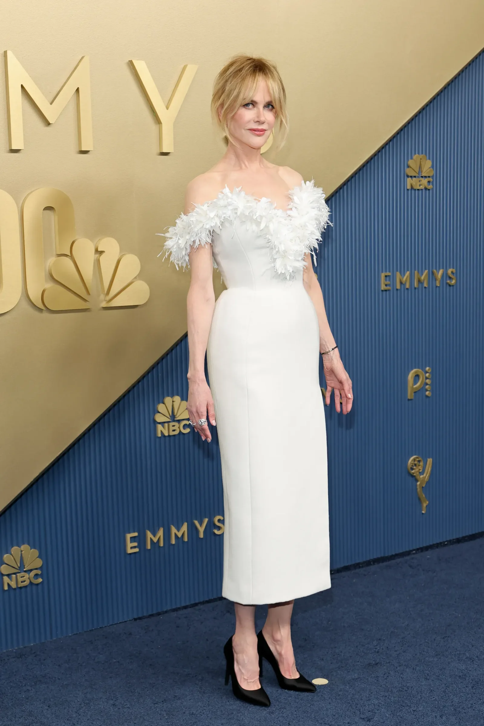 Woman in a white gown with feather-embellished neckline posing on a blue-and-gold step-and-repeat backdrop at the Emmys.
