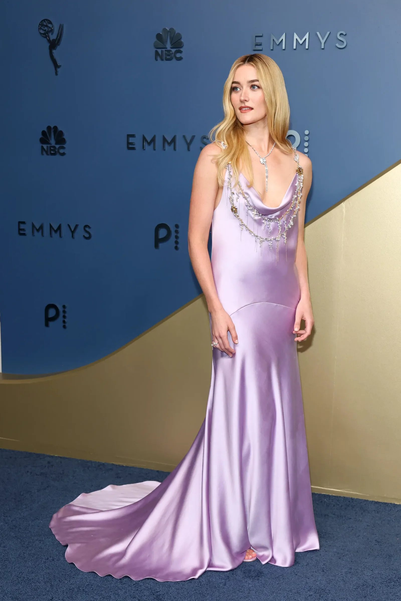 Full-length view of a woman in a lavender satin gown with a bejeweled necklace posing on a blue Emmy Awards backdrop.