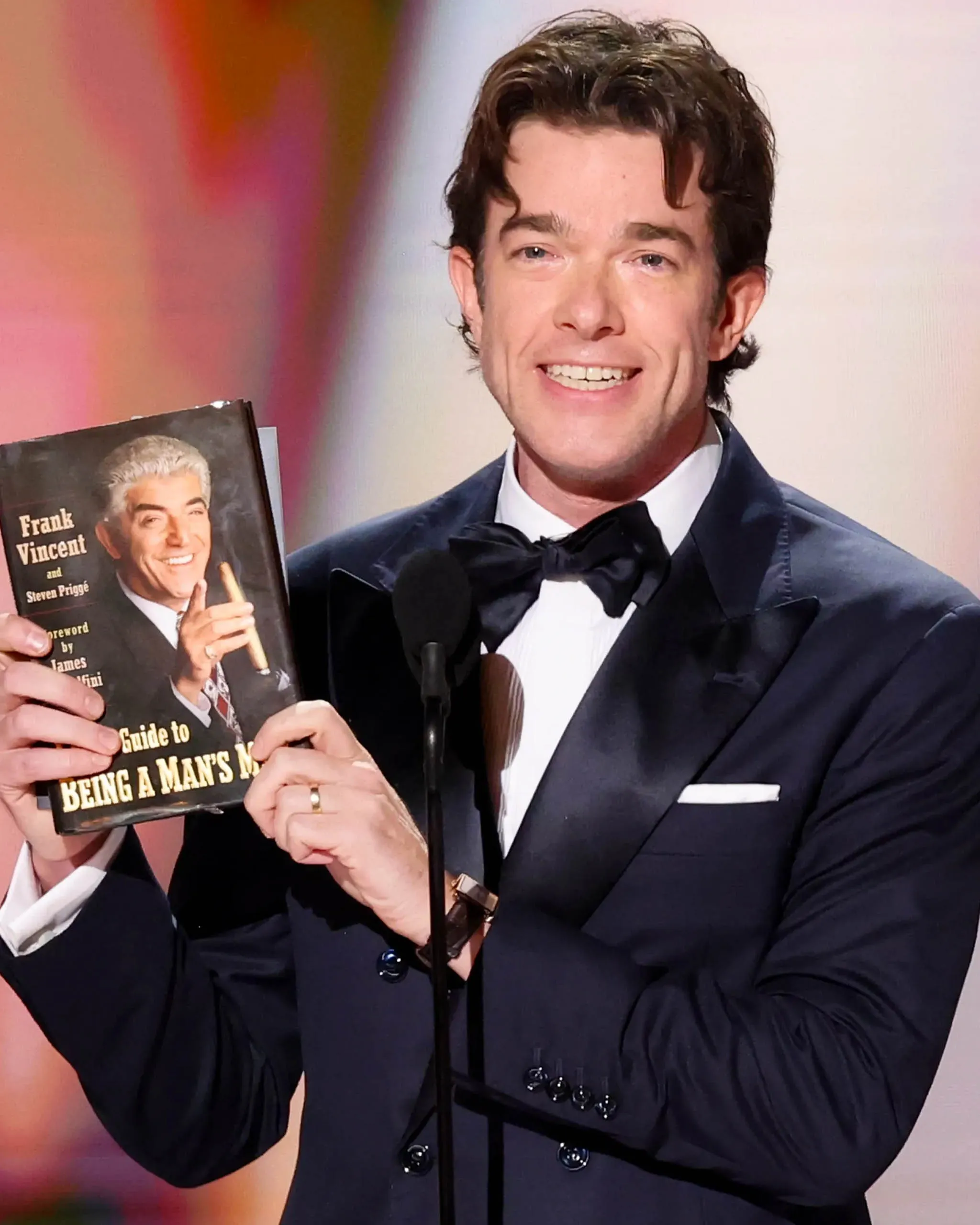 Smiling man in a navy tuxedo holds up a book on stage, standing at a microphone.