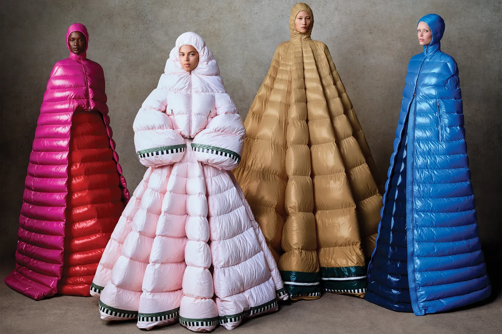 Four models pose in floor-length, oversized puffer coats in pink, white, beige, and blue against a gray backdrop.