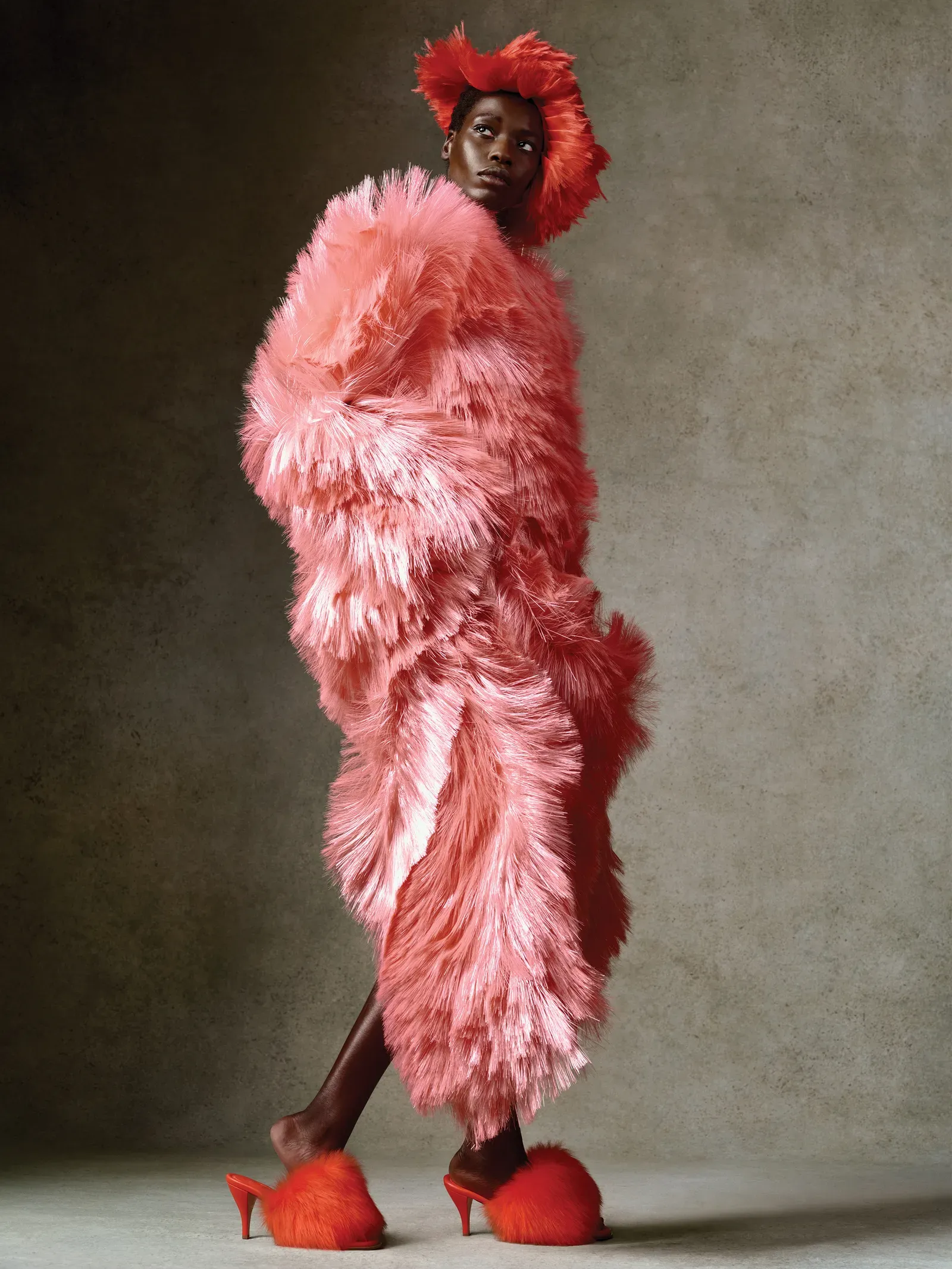 Model in a voluminous pink feather coat with a red feathered headpiece, wearing red high heels, posed against a textured gray backdrop.