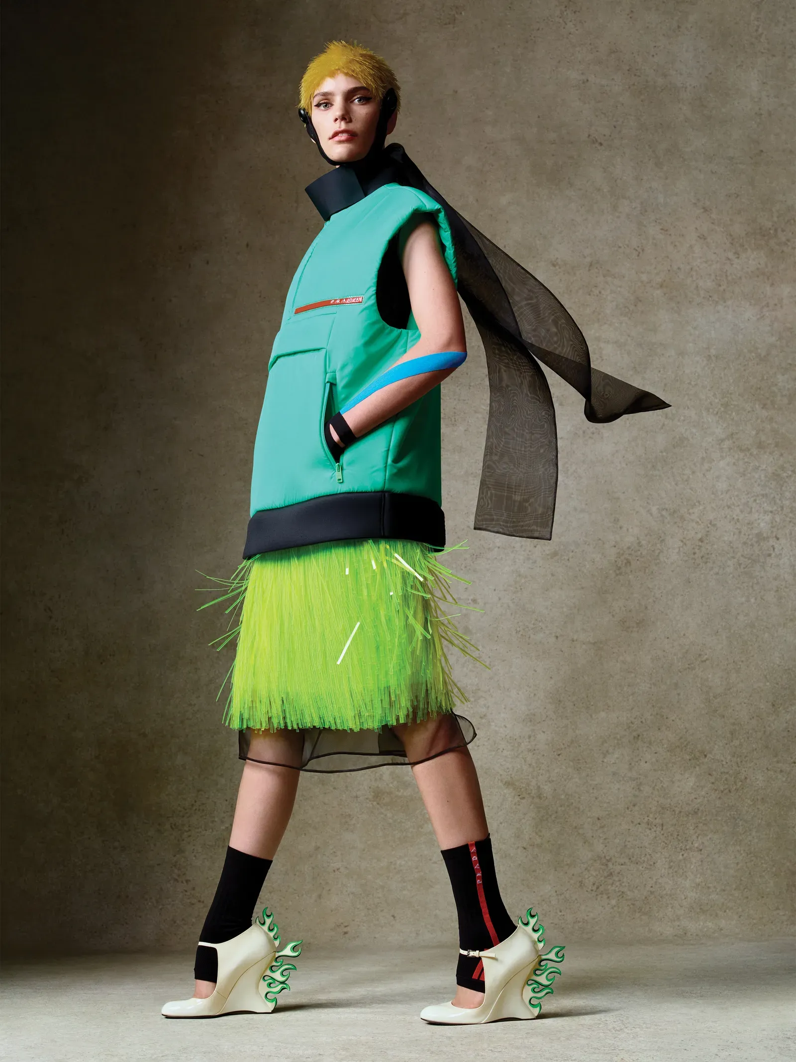 Model wearing a bright turquoise padded vest with a neon fringe skirt, and a black flowing cape, finished with cream boots featuring green flame accents.