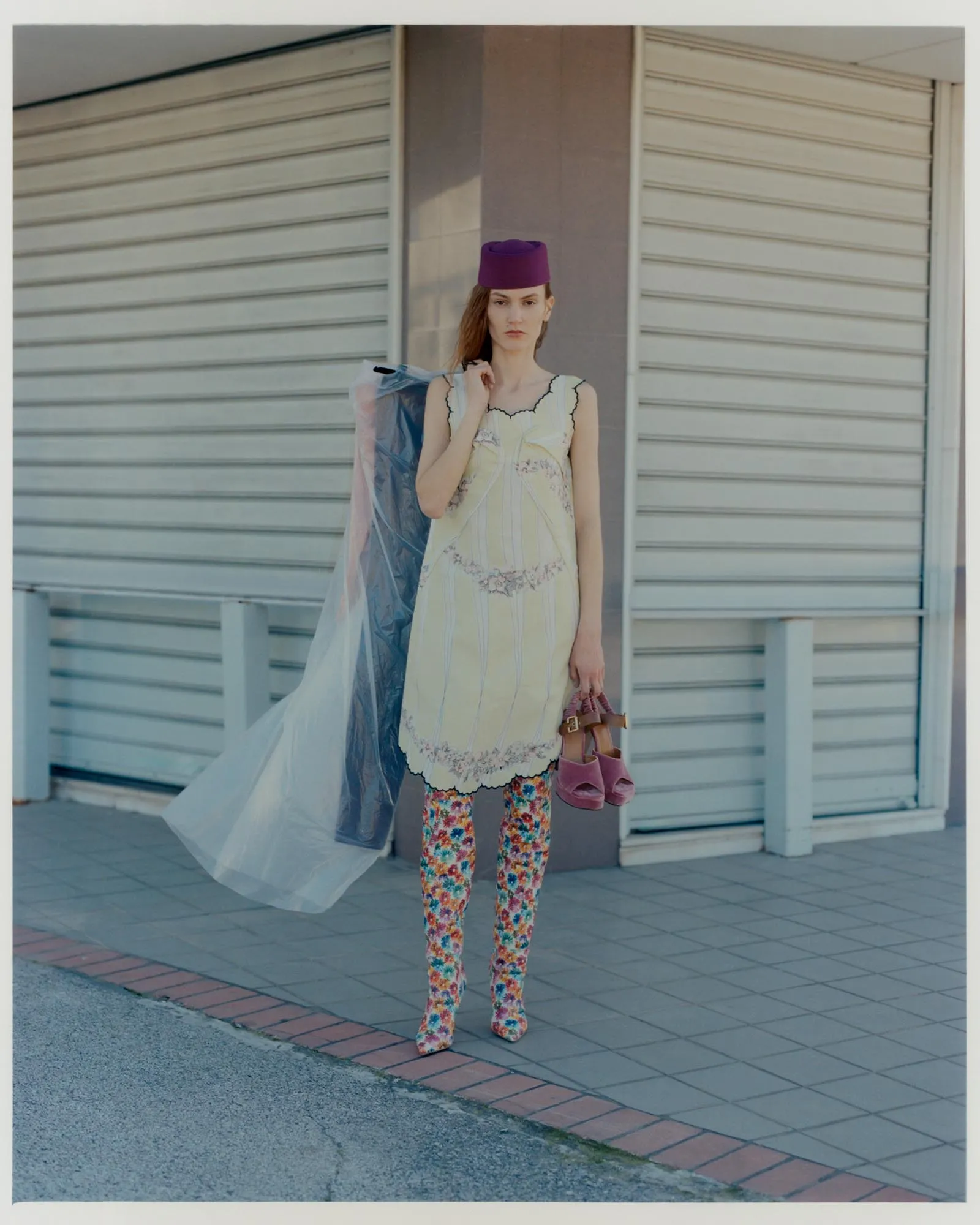 Woman standing on a sidewalk in front of closed shop shutters, wearing a pale dress with floral tights, a purple hat, a sheer cape, and carrying a pink handbag.