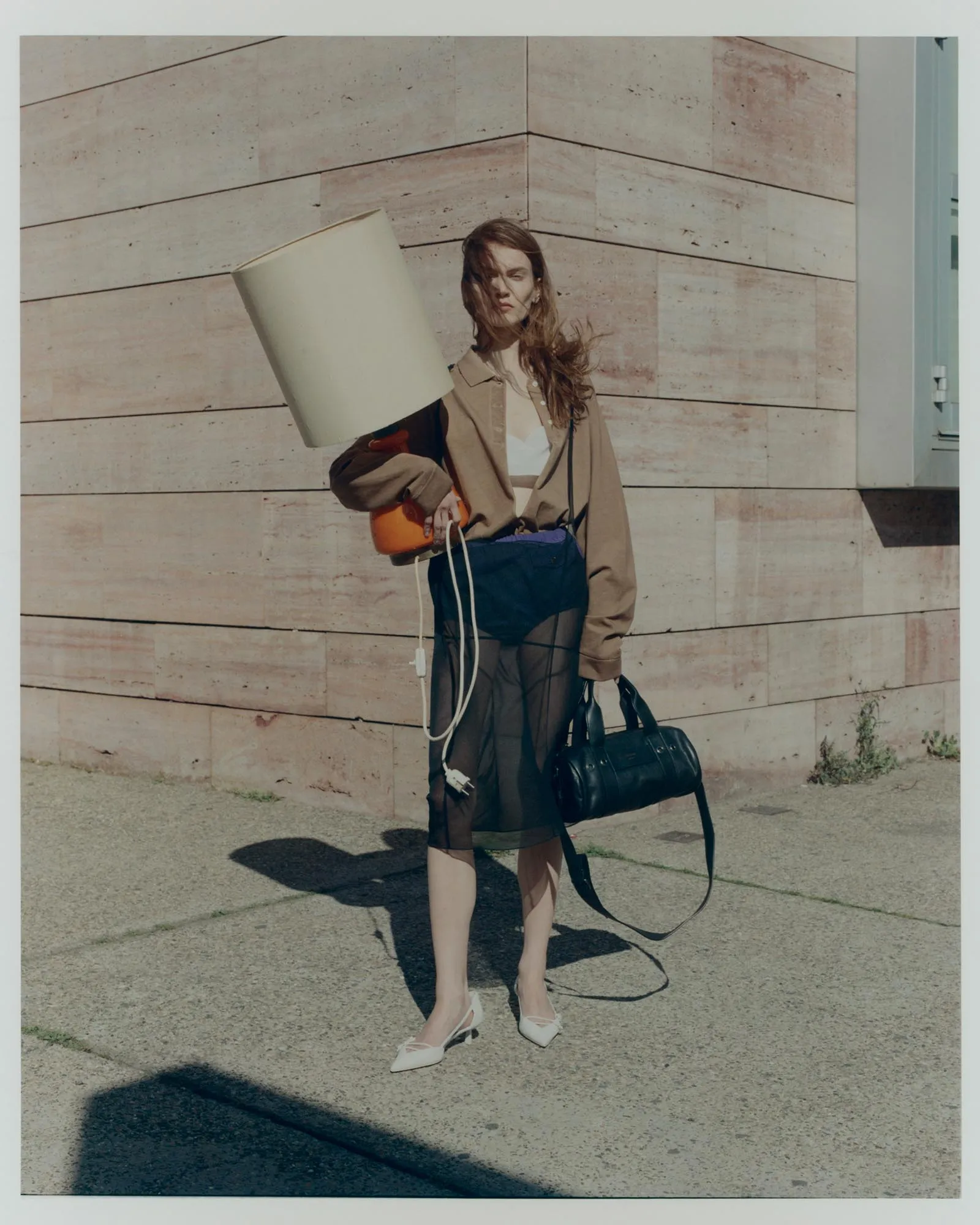 Model posing in a beige trench and white top, holding a large lampshade on her shoulder outdoors against a stone wall.