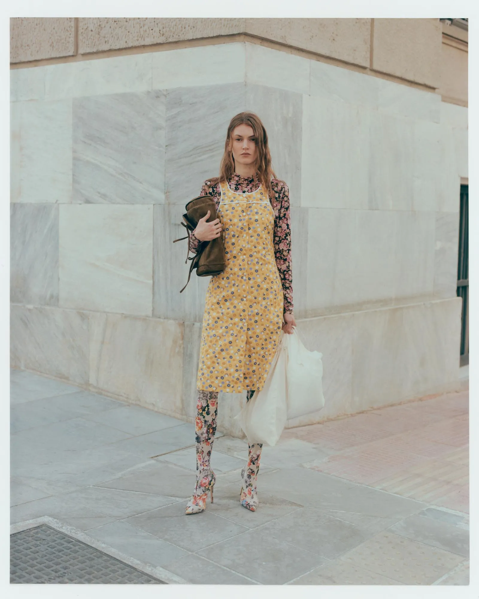 Young woman stands against a light marble wall, wearing a yellow floral dress over a floral top and carrying a brown backpack.