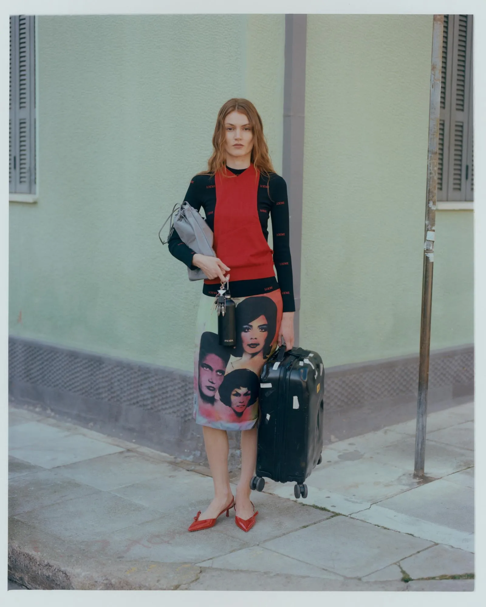 Woman standing on a sidewalk with a rolling suitcase and a gray handbag, wearing a red dress over a black long-sleeve top and a skirt with colorful face prints against a pale wall.