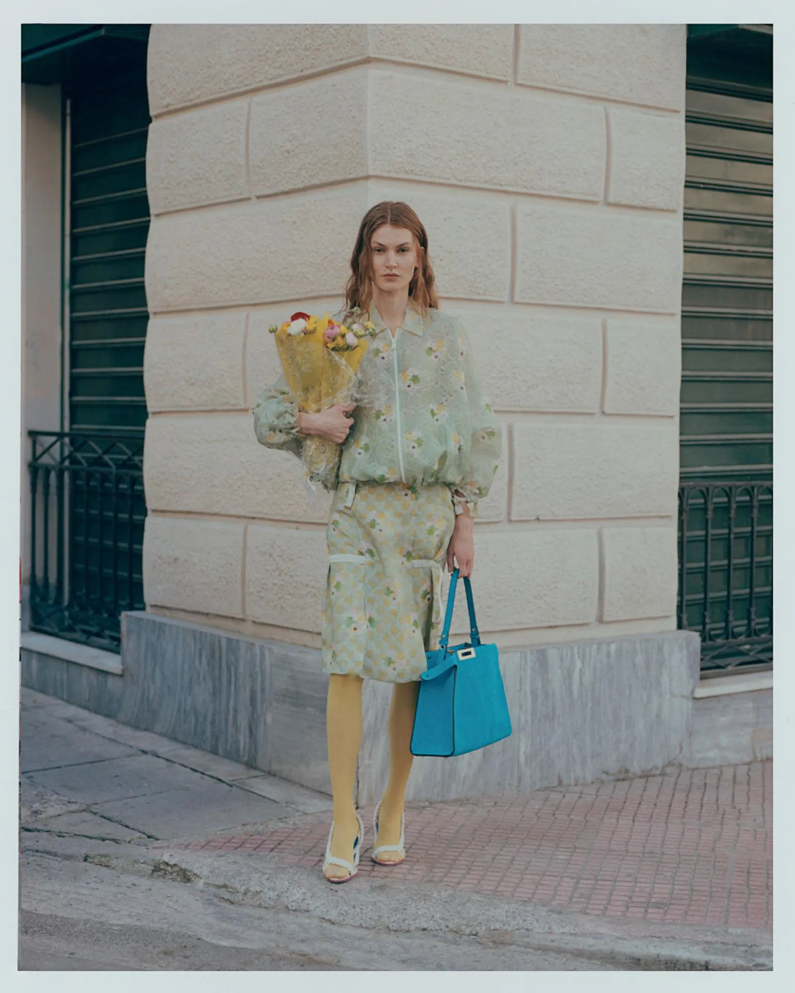 Woman standing on a city sidewalk holding a bouquet of flowers in yellow wrapping, wearing a pastel floral outfit and blue handbag against a stone building backdrop.