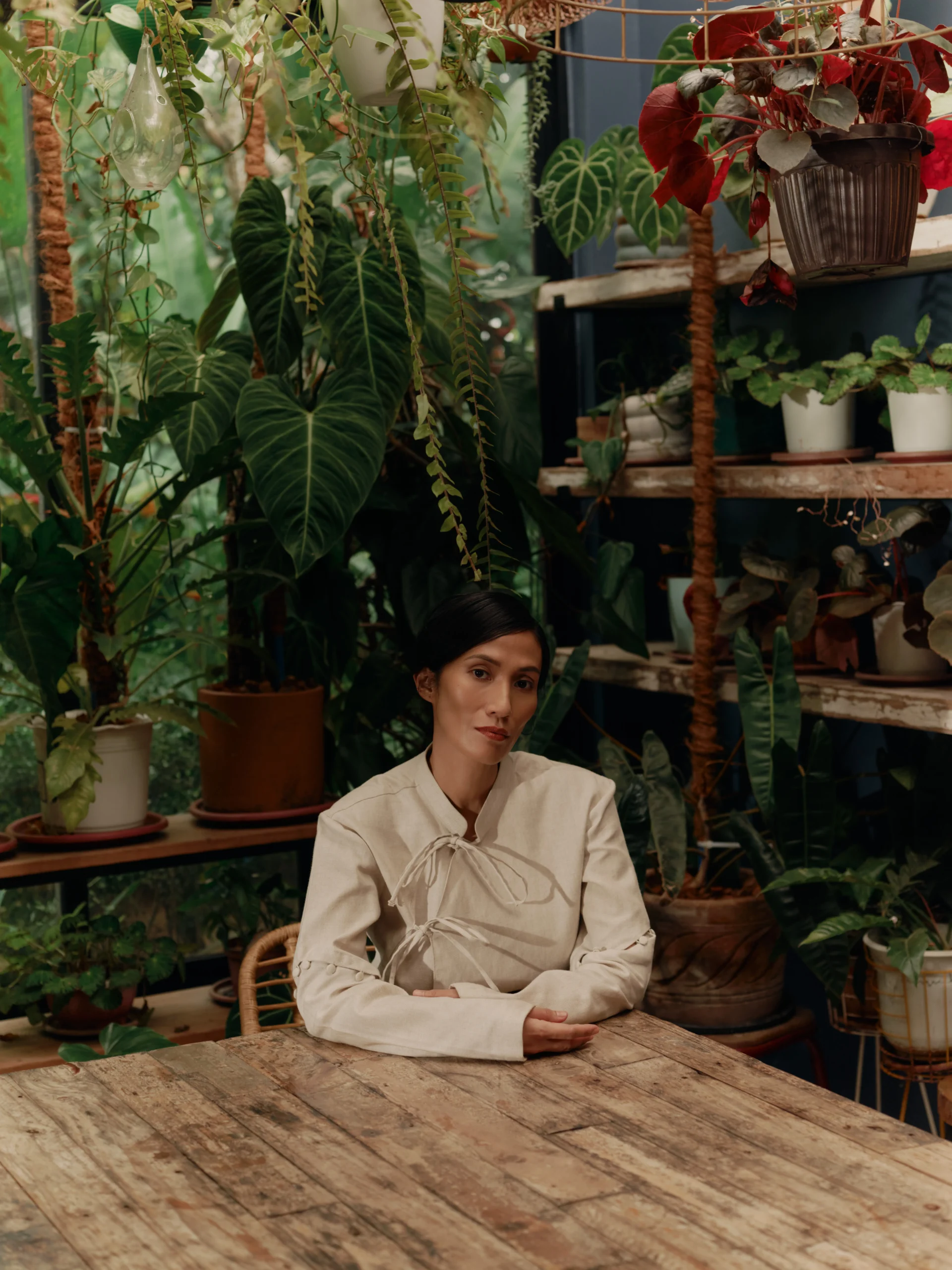 Woman seated at a rustic wooden table in a greenhouse filled with hanging and potted plants.