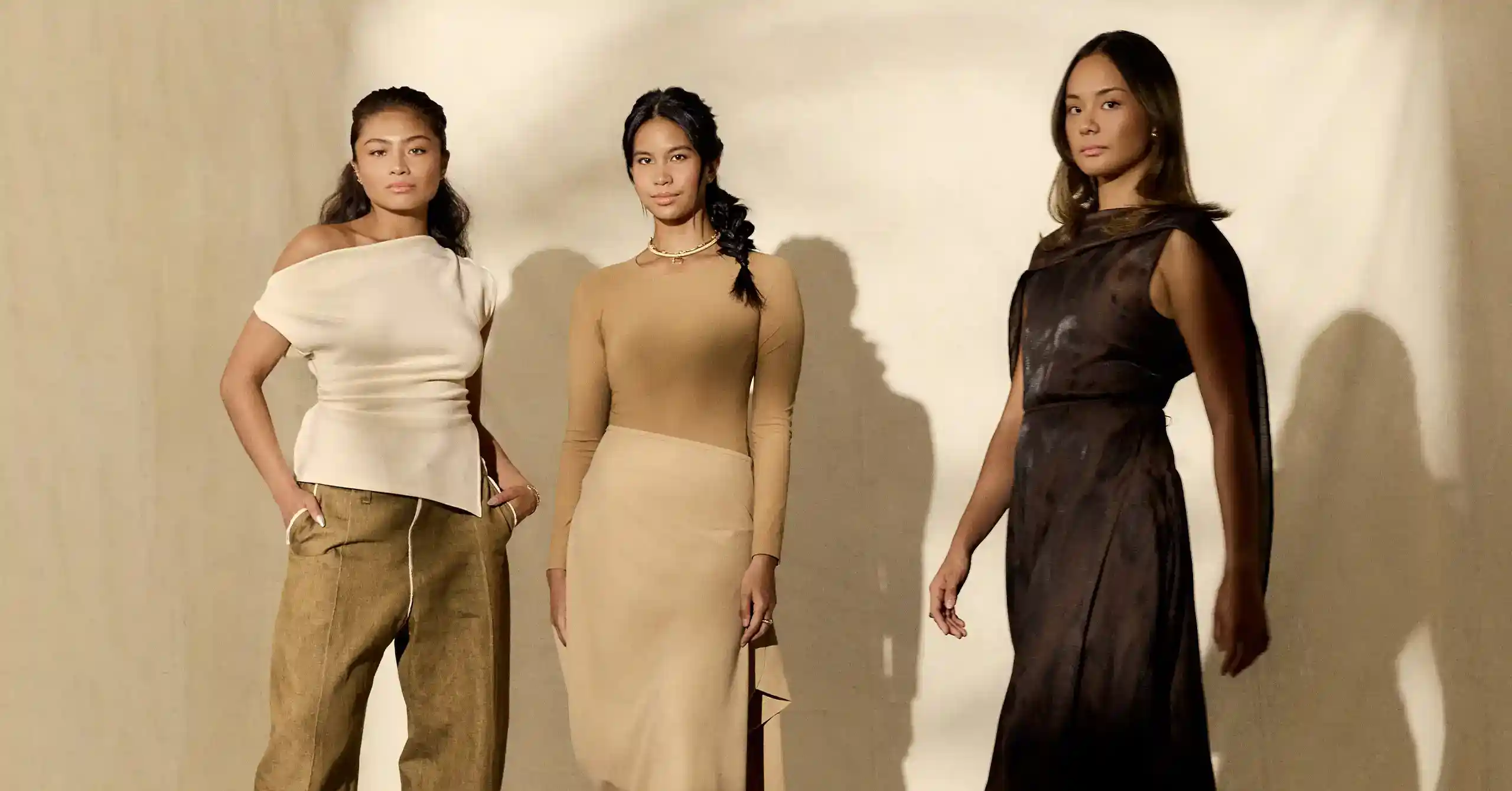 Three women pose in a beige-toned fashion shoot, wearing elegant dresses: white off-shoulder, cream long-sleeve, and dark brown gown against a light backdrop.