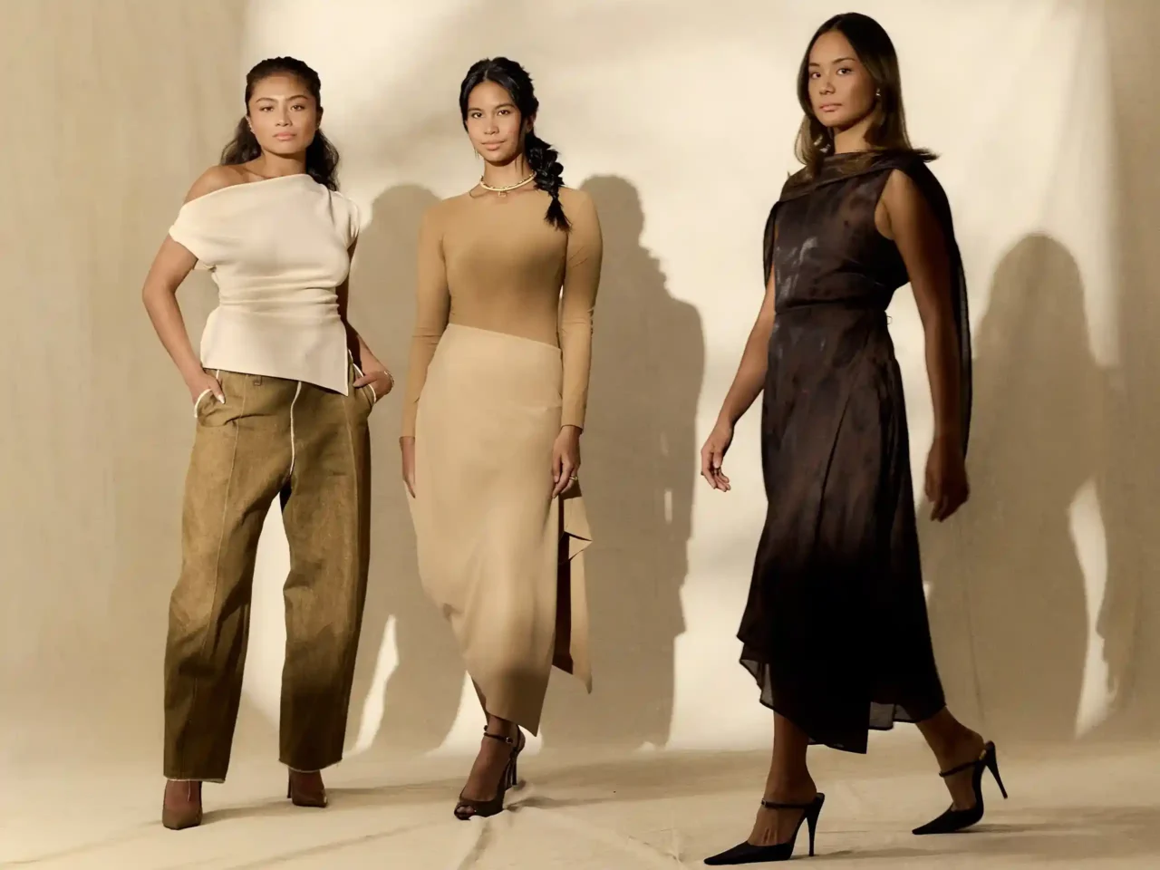 Three women pose in a beige-toned fashion shoot, wearing elegant dresses: white off-shoulder, cream long-sleeve, and dark brown gown against a light backdrop.