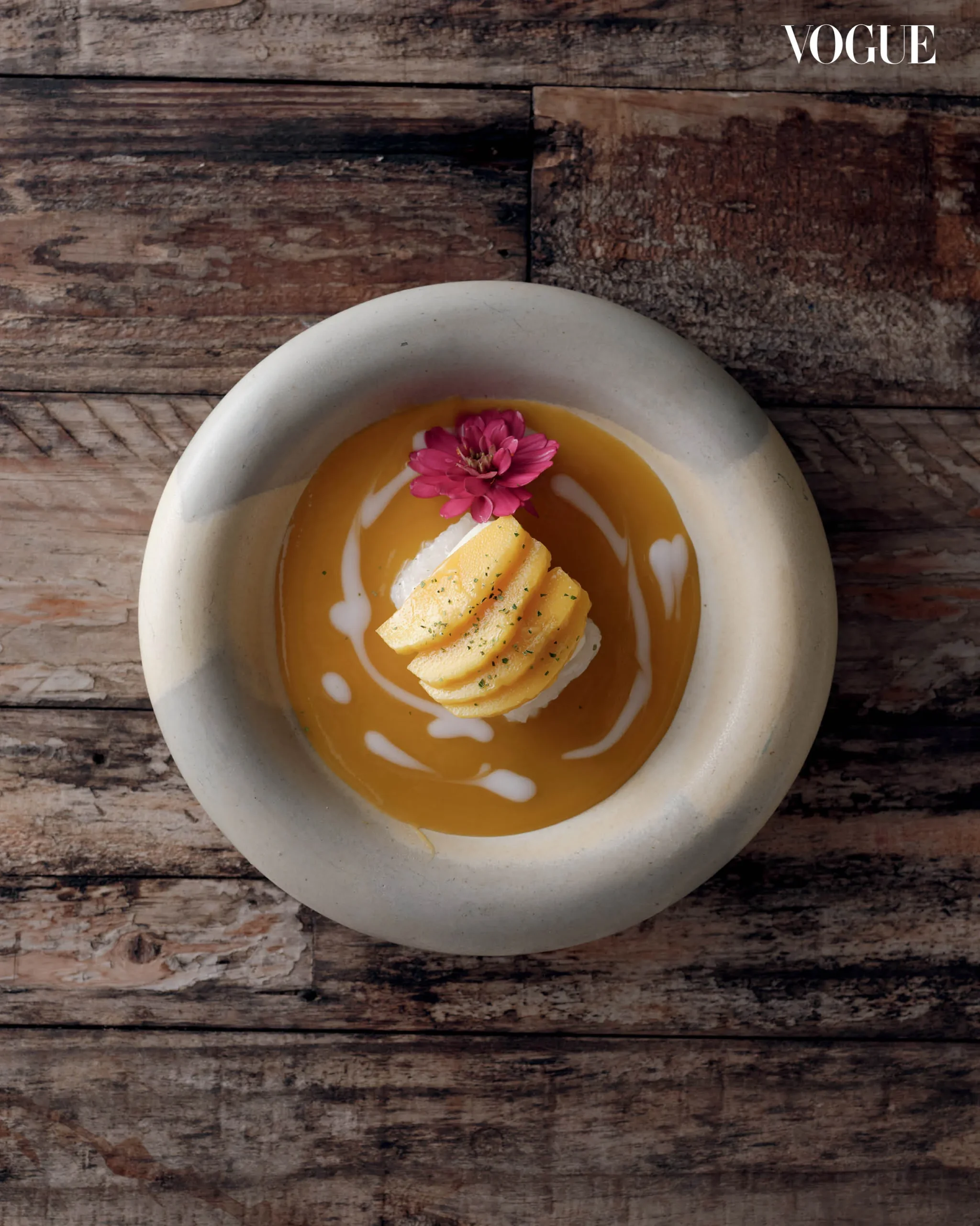 Top-down view of orange soup in a ceramic bowl, garnished with mango slices and a pink flower on a rustic wooden table; Vogue watermark in corner.