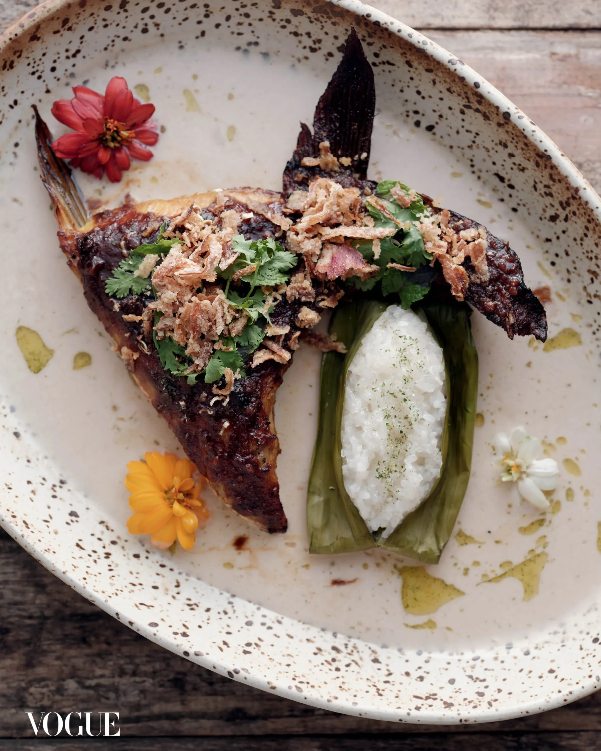 Roasted fish fillet with crispy fried onions and fresh cilantro, served with a white sauce and green vegetables on a speckled plate, garnished with orange flowers.