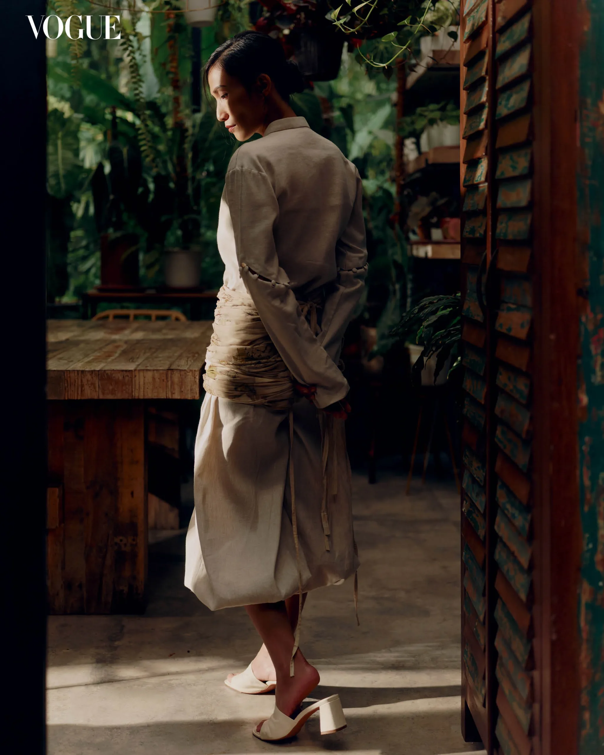 Side view of a woman in a beige outfit walking through a plant-filled space with a Vogue logo in the corner.
