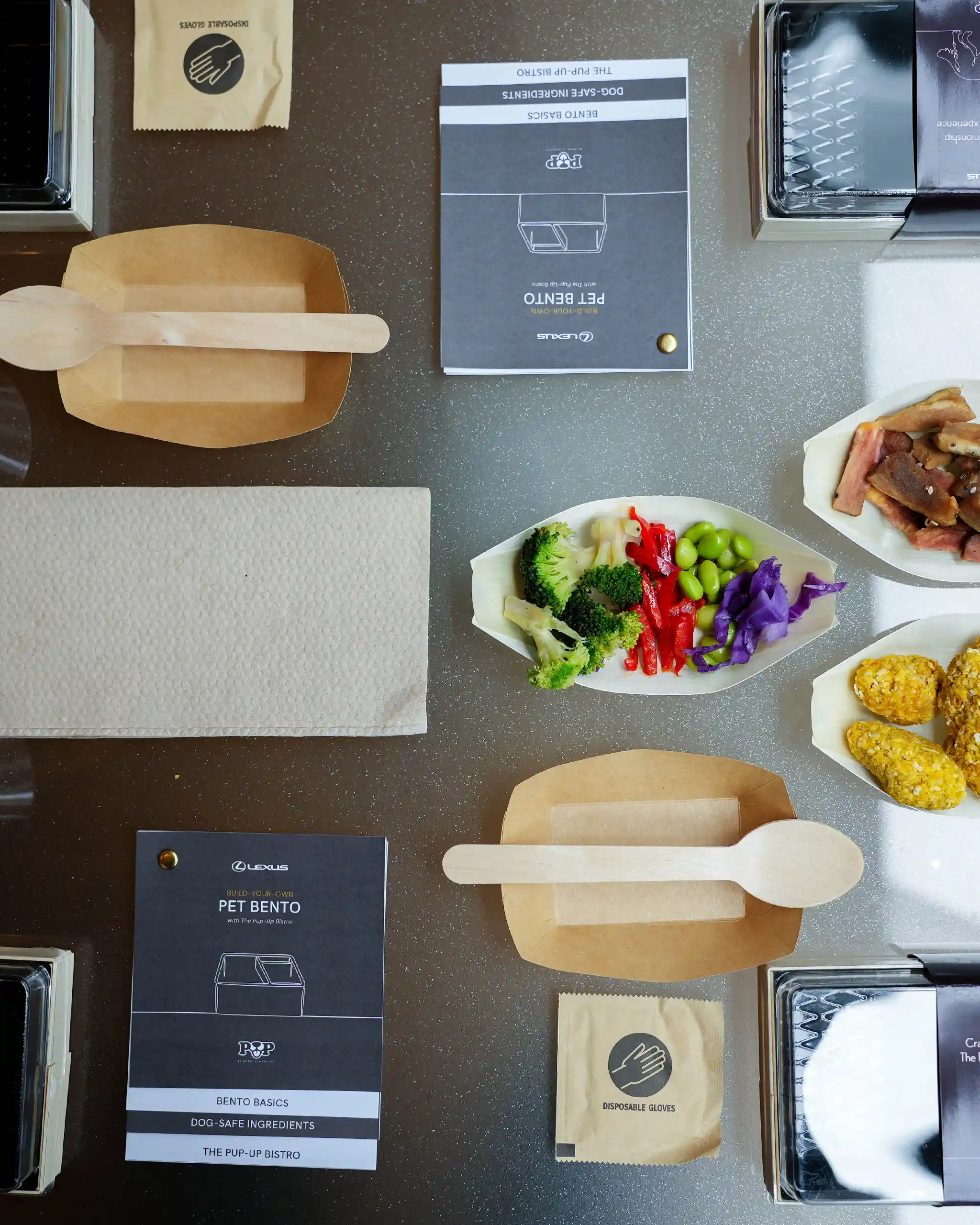 Top-down view of a pet-bento kit on a speckled counter, showing a recipe booklet, disposable gloves, wooden utensils, and portions of colorful vegetables and cornmeal treats in trays.