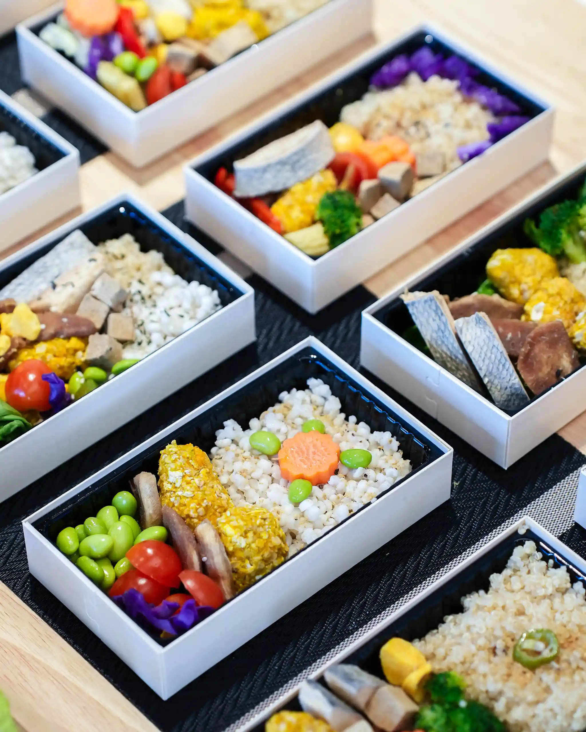 Row of white bento boxes filled with rice topped with colorful veggies (carrot flower, edamame) and side components, arranged on a dark mat.