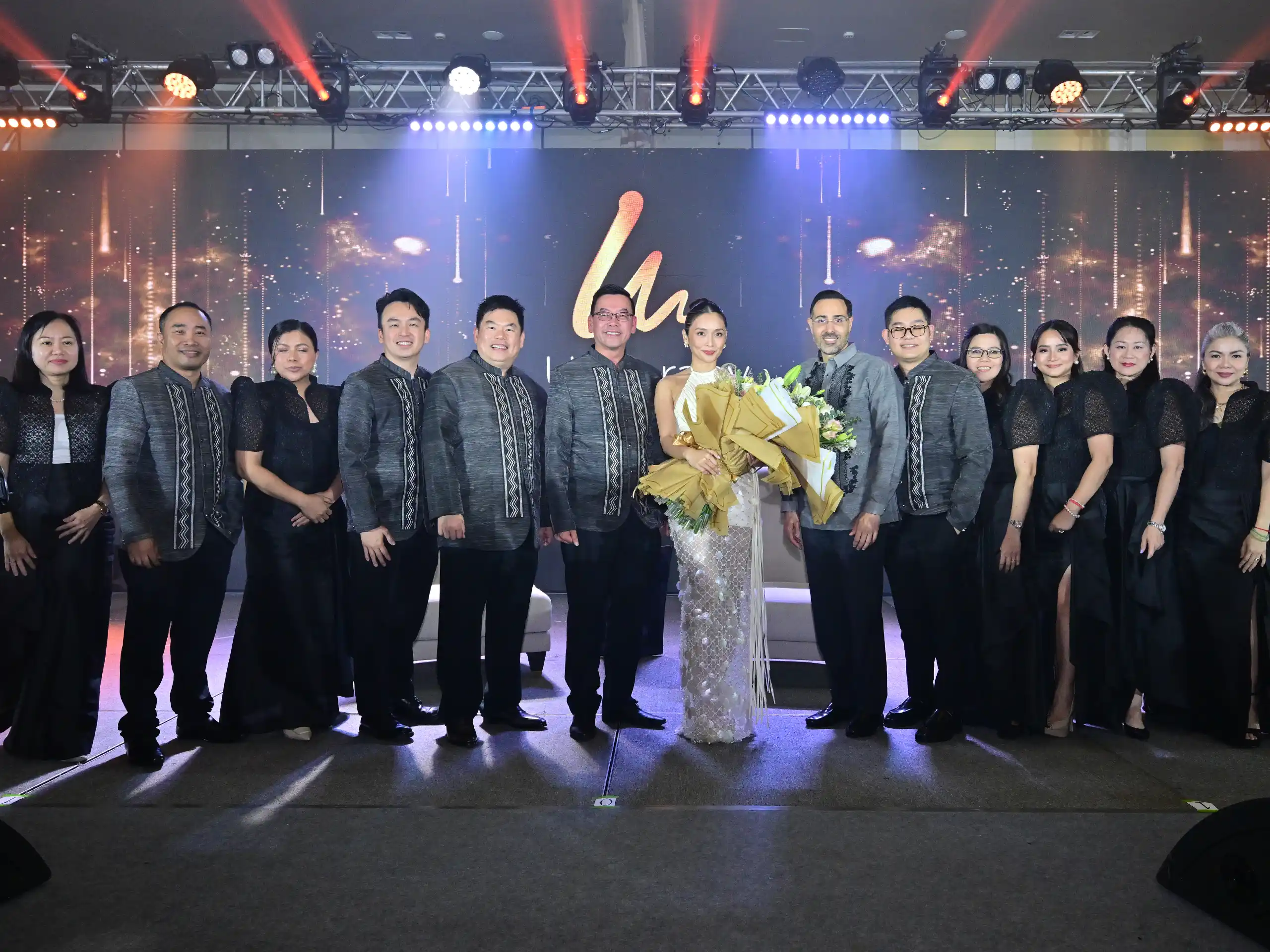 Group of people in matching gray outfits posing on a stage, center woman holding bouquets of flowers.