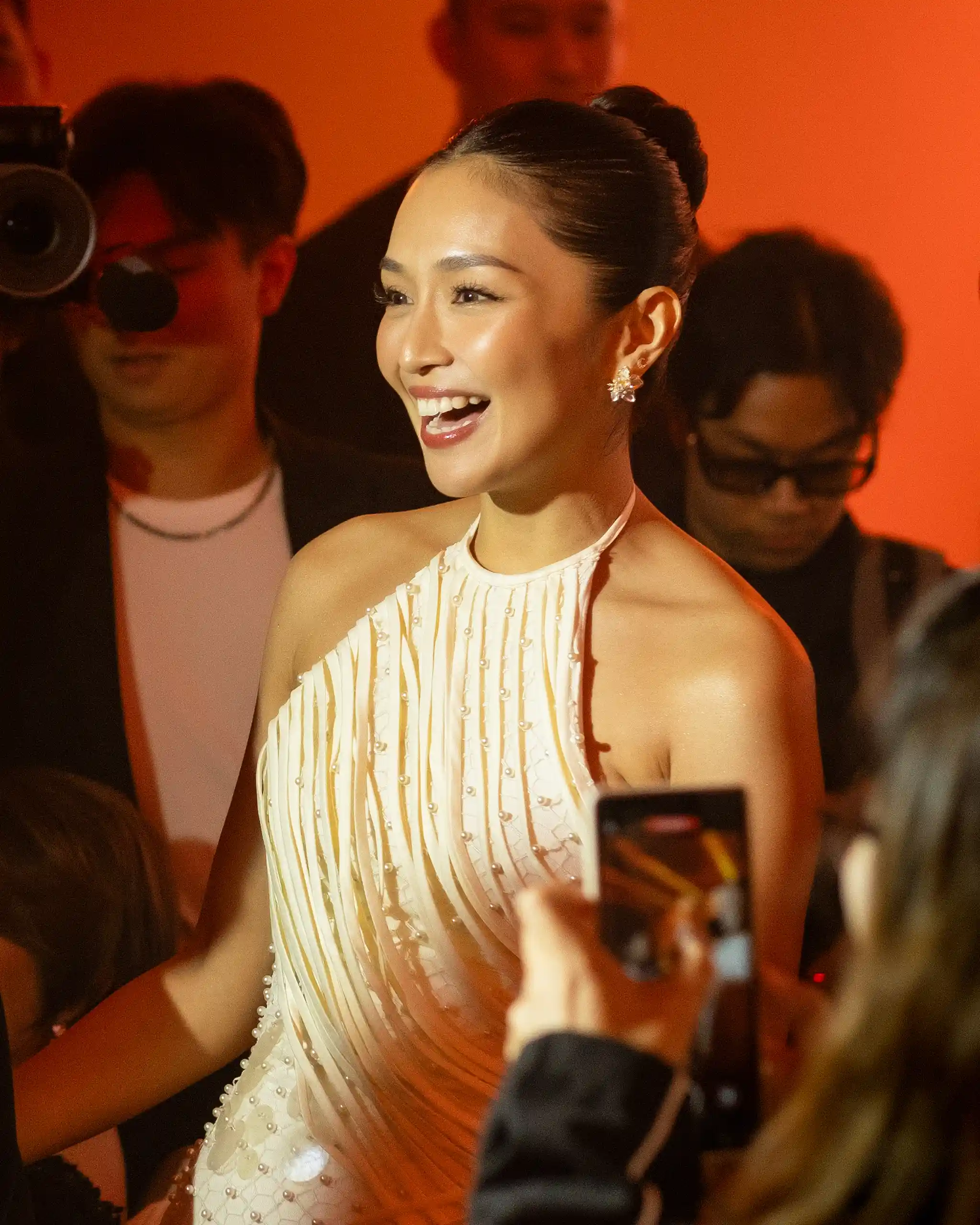 Smiling woman in a cream beaded halter dress at a red-lit event, surrounded by photographers.