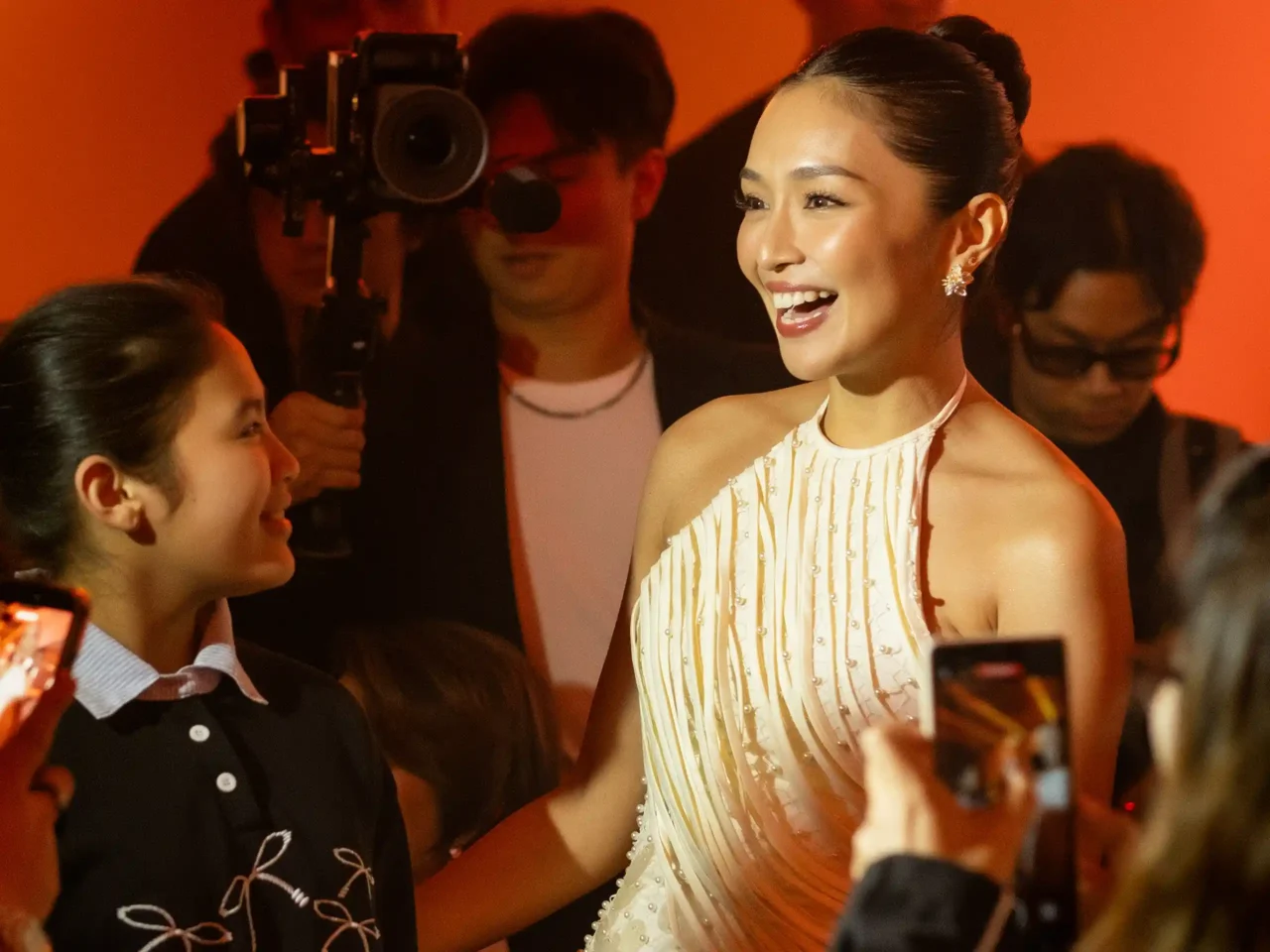 Smiling woman in a pale cream sequined dress chats with fans at a crowded event; orange lighting and photographers visible in the background.