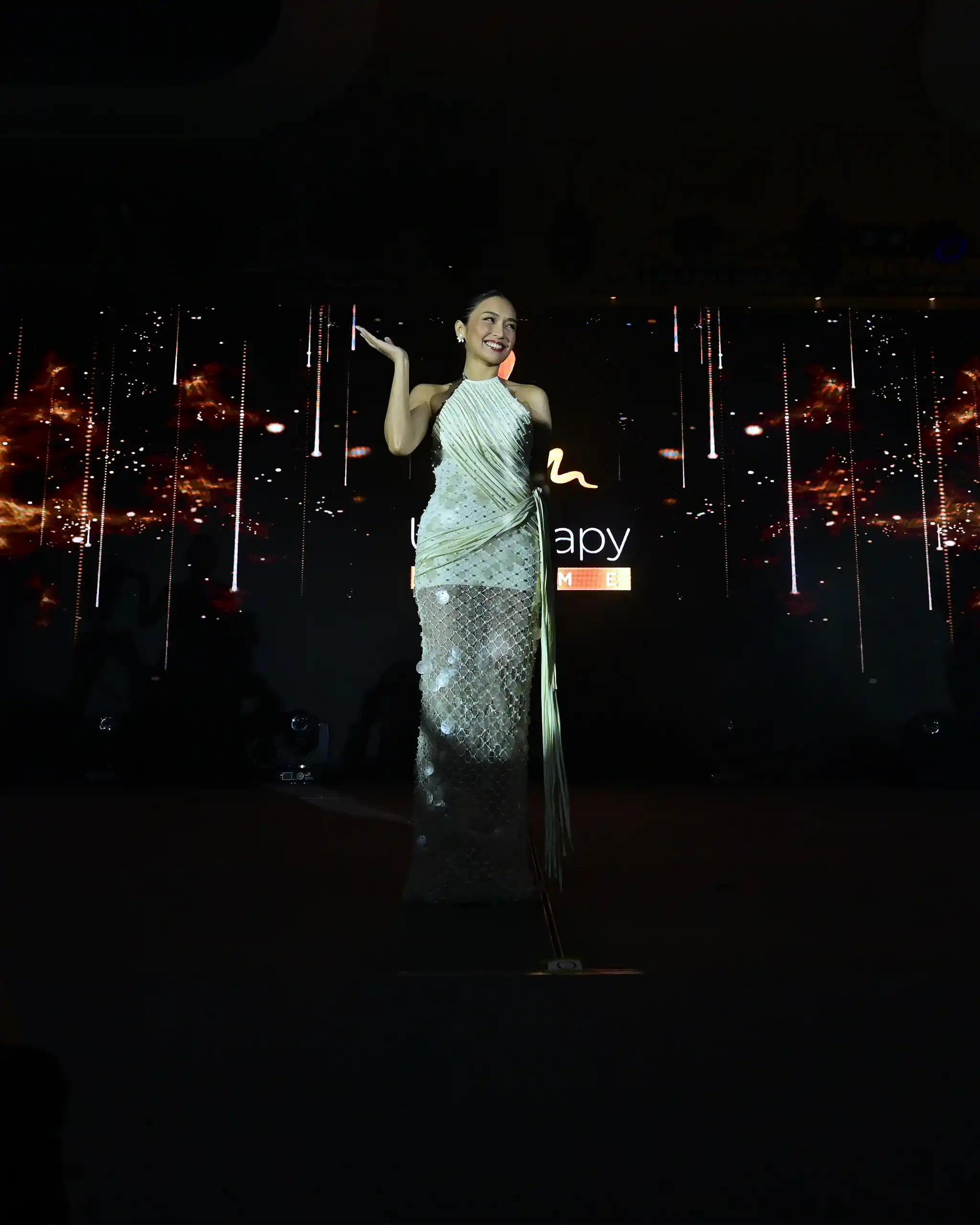 Smiling female performer in a silver, beaded gown on a dark stage with her hand raised and colorful lights behind her.