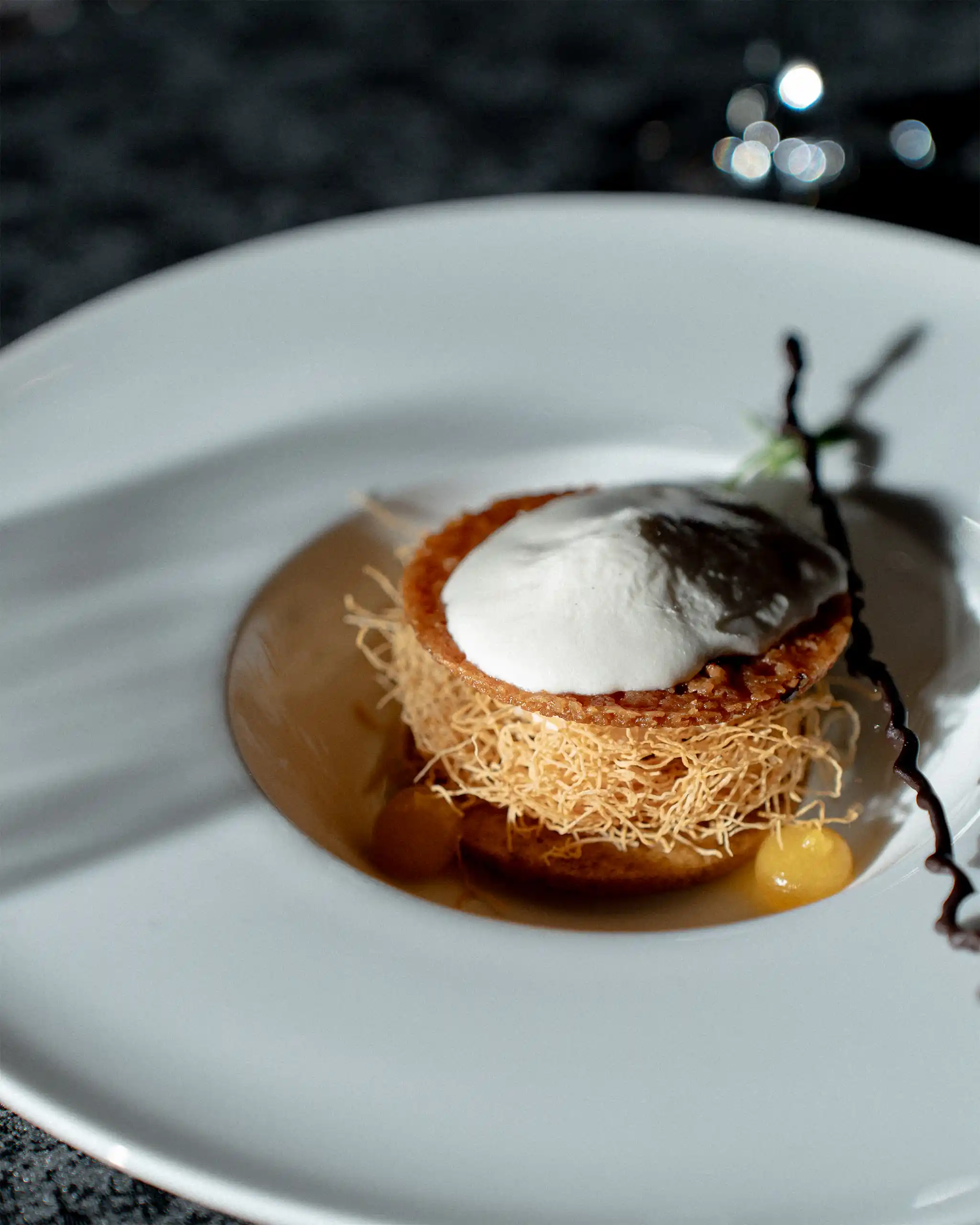 Close-up of a circular dessert with crisp shredded pastry and a dollop of white cream on top, served on a white plate against a dark, blurred background.