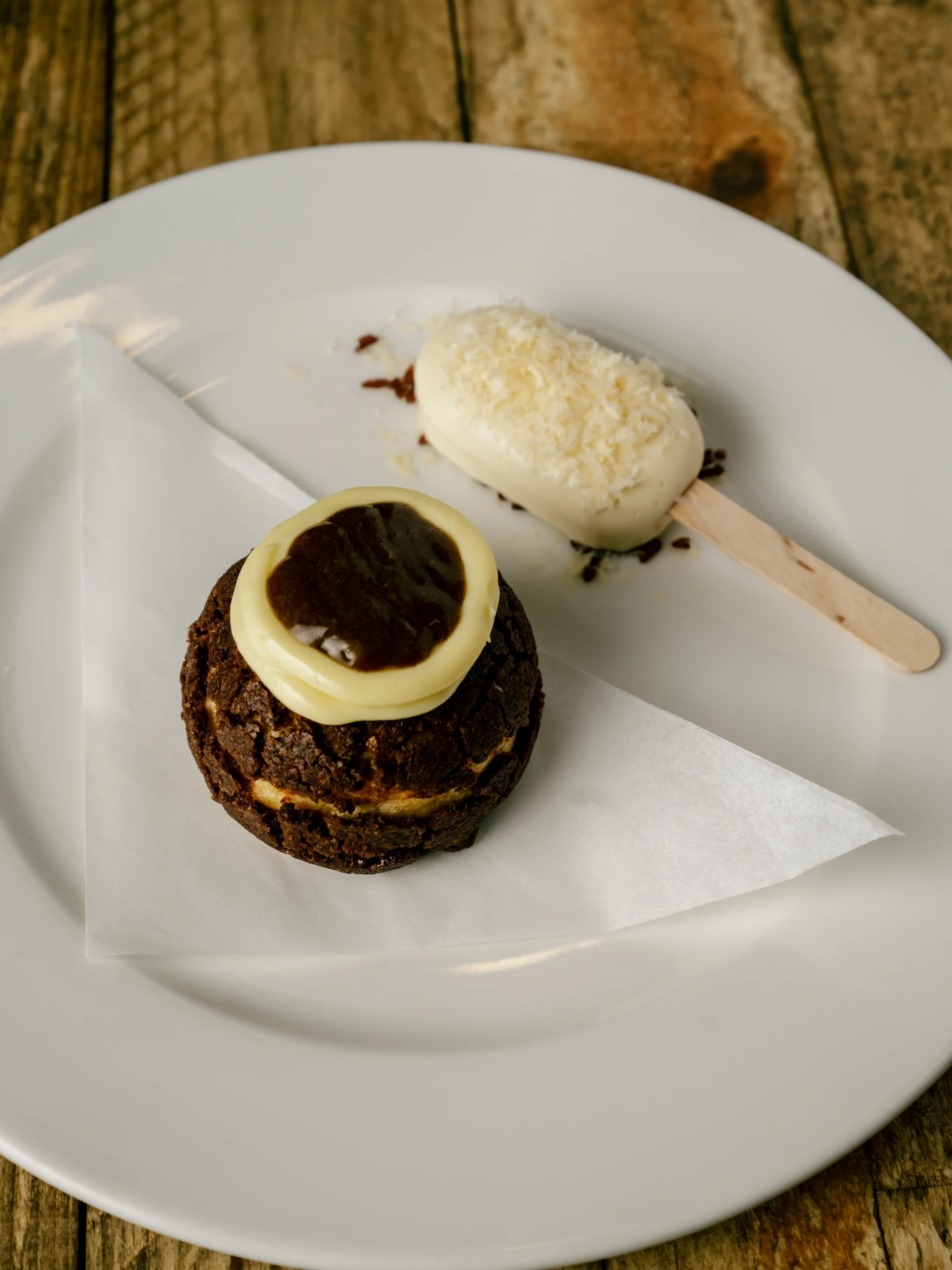 Chocolate cupcake with a white chocolate garnish on a white plate; beside it a coconut-dusted vanilla ice cream bar on a wooden stick.
