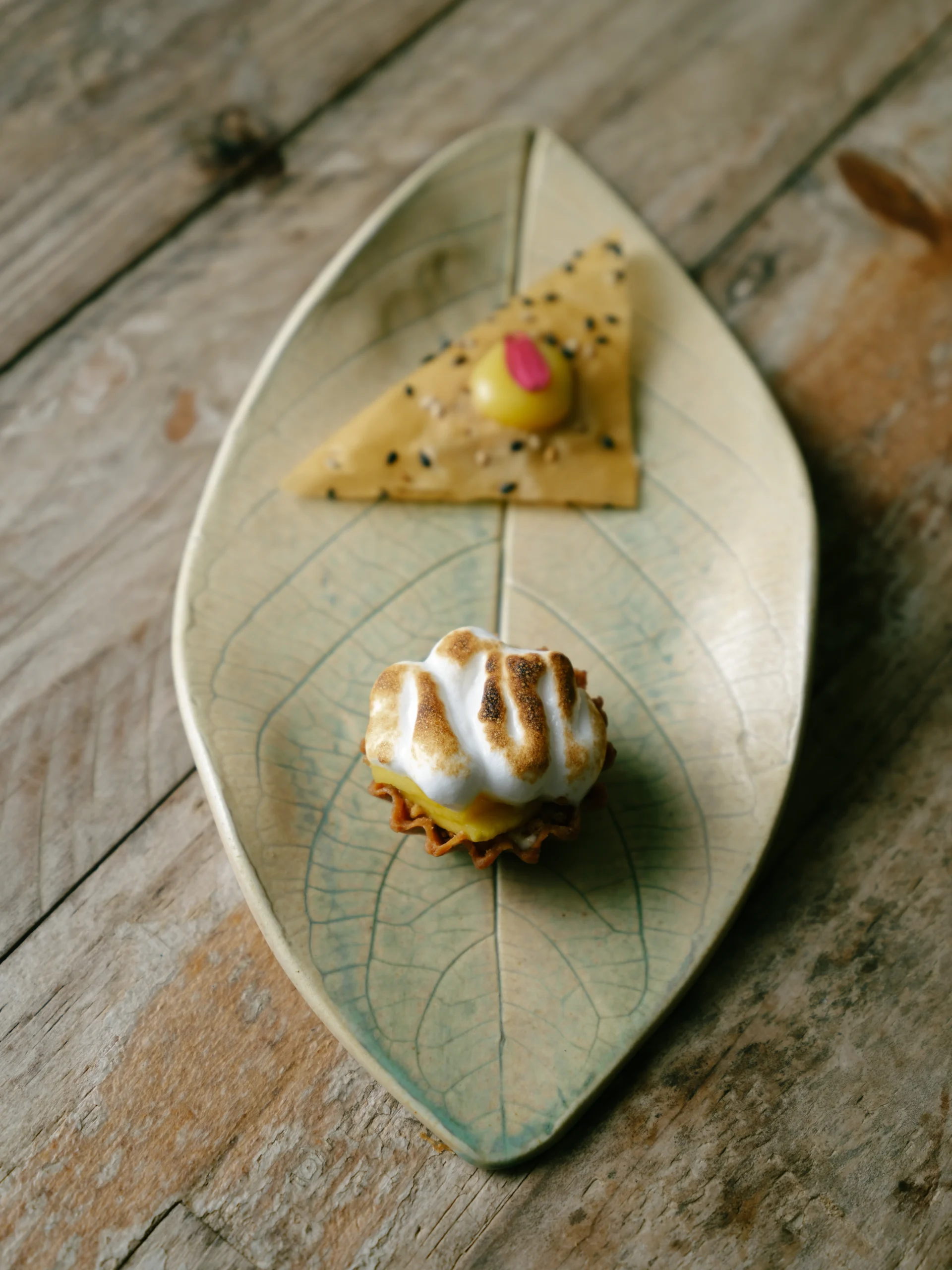 Leaf-shaped plate on a wooden table holds a sesame-crusted triangular cracker with a yellow olive bite and a toasted meringue-topped dessert.