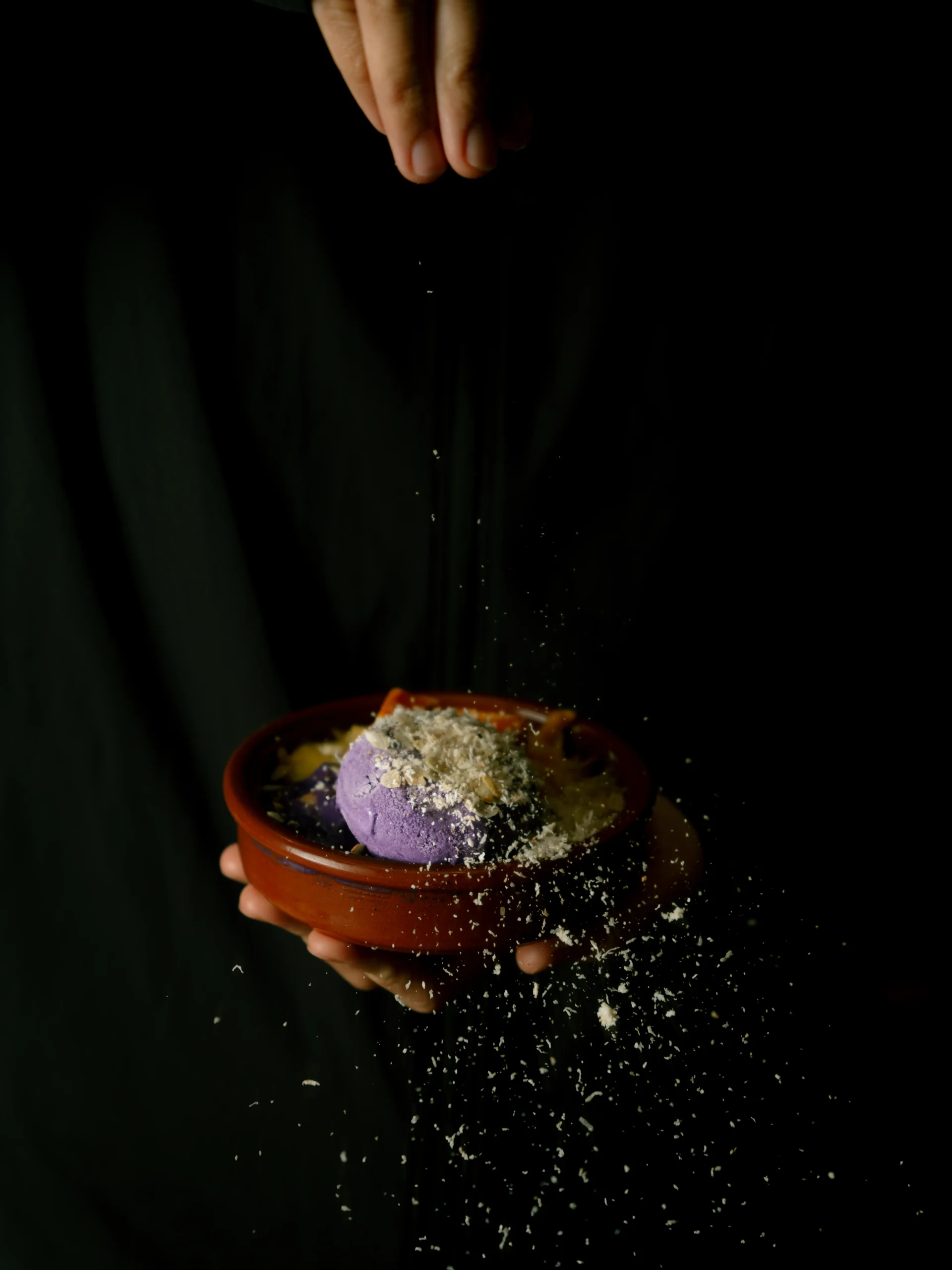 Hands hold a terracotta bowl with two purple ice cream scoops as white crumbs scatter down against a dark background.