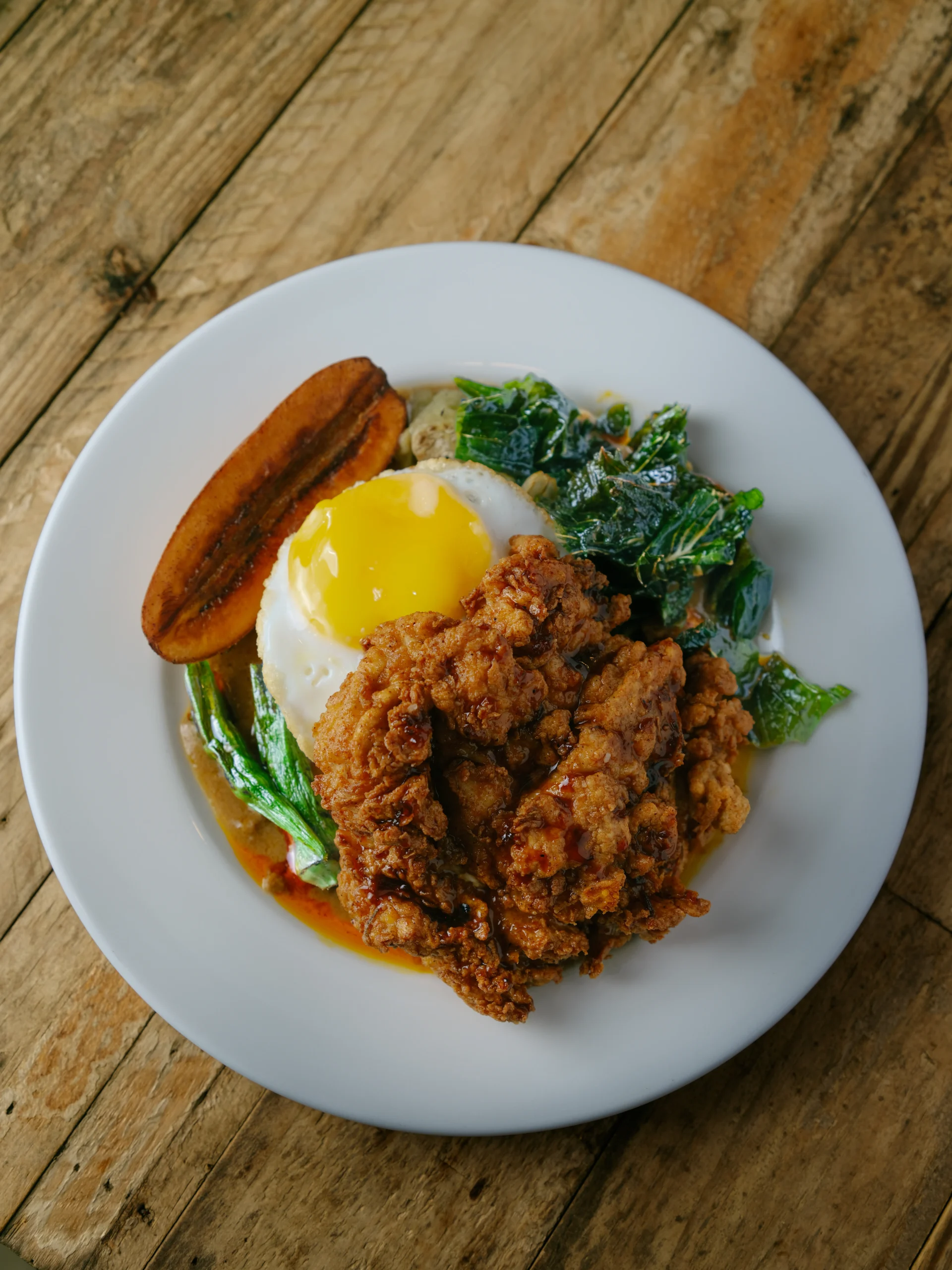 Plate with fried chicken, sunny-side-up egg, sautéed greens, and a fried plantain on a rustic wooden table.