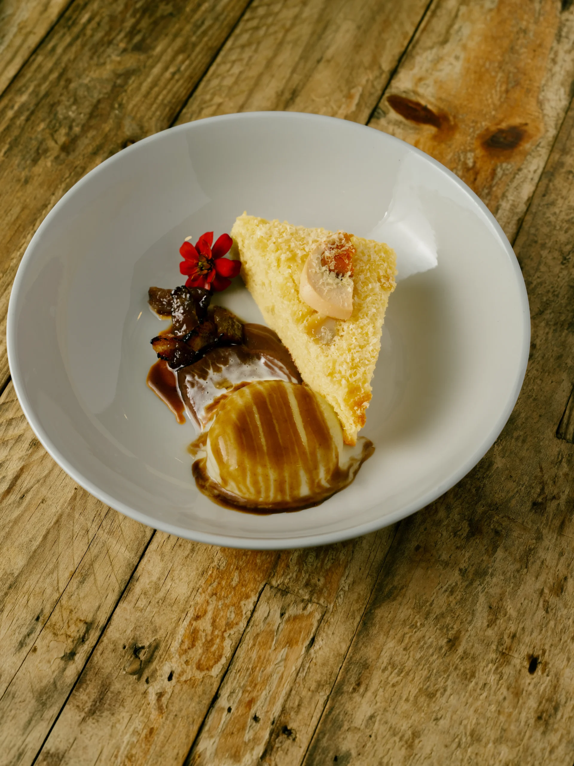 Slice of yellow cake with cream topping on a white plate, accompanied by a red flower garnish and caramel sauce on a rustic wooden table.