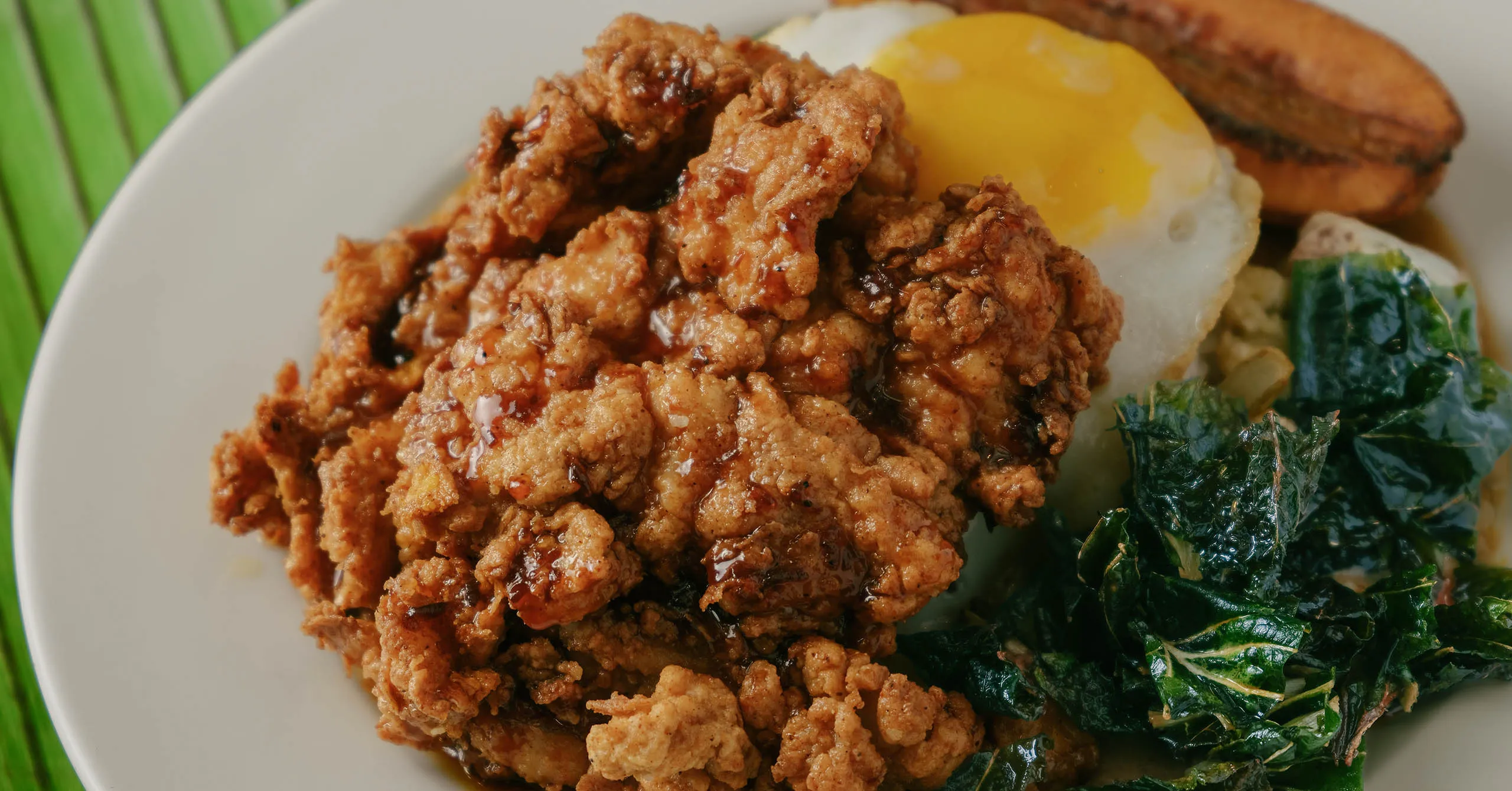 Crispy fried chicken on a plate with a sunny-side-up egg, sautéed kale, and plantain slices in the background.