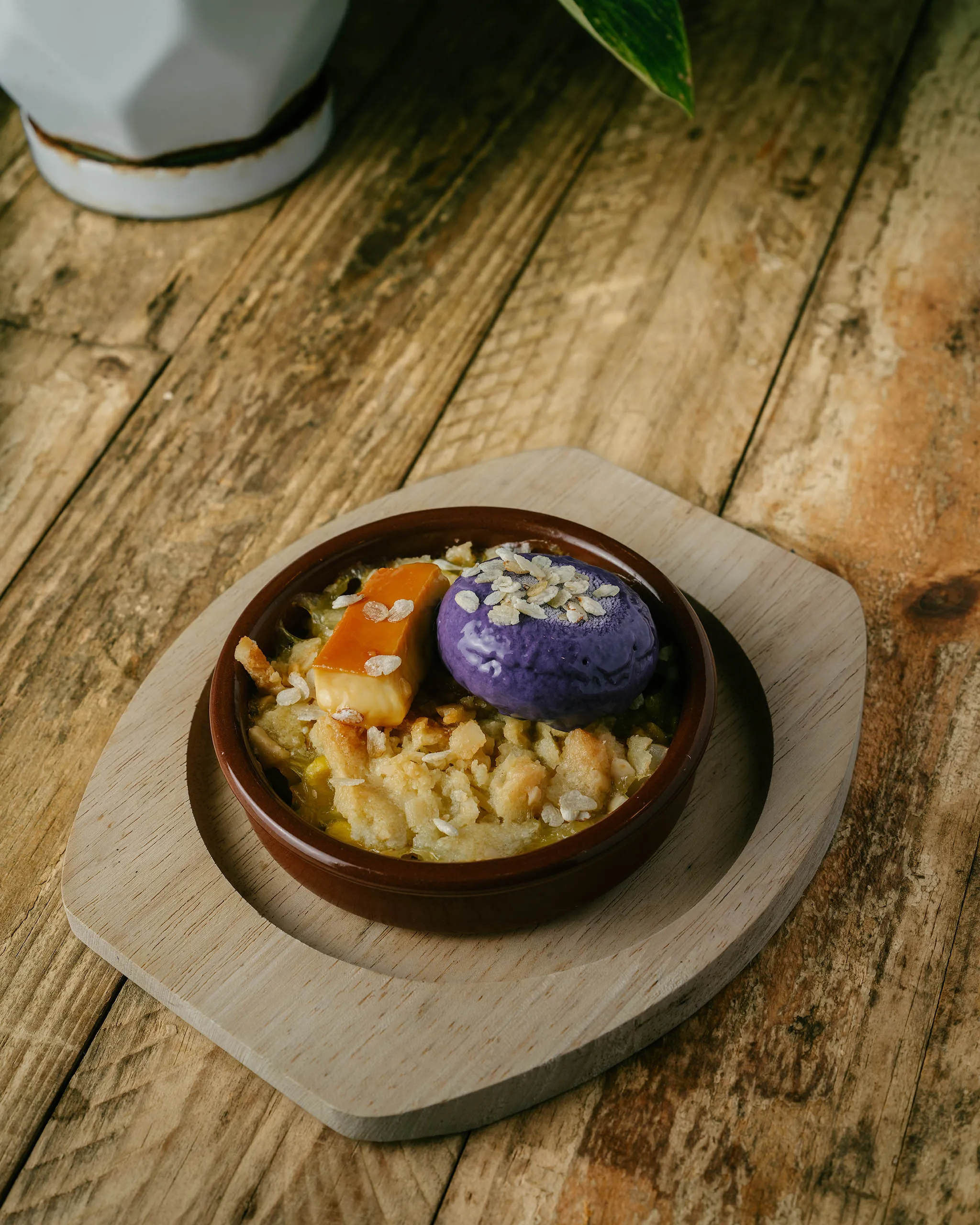 Brown ceramic dessert bowl on a wooden board with purple ice cream scoop, oat toppings, and diced fruit or nuts, on a rustic table.