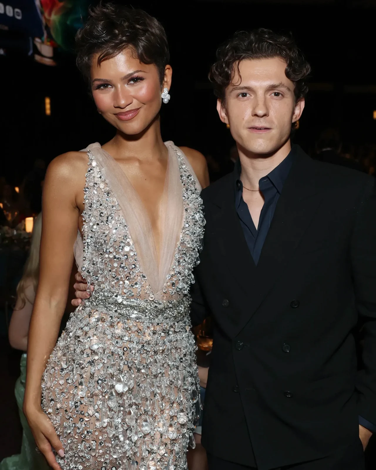Close-up of a smiling woman and man posing at a formal event, woman wearing a beaded gown and large drop earrings, man in a dark suit against a dark background.