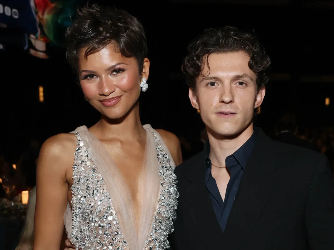 Close-up of a smiling woman and man posing at a formal event, woman wearing a beaded gown and large drop earrings, man in a dark suit against a dark background.