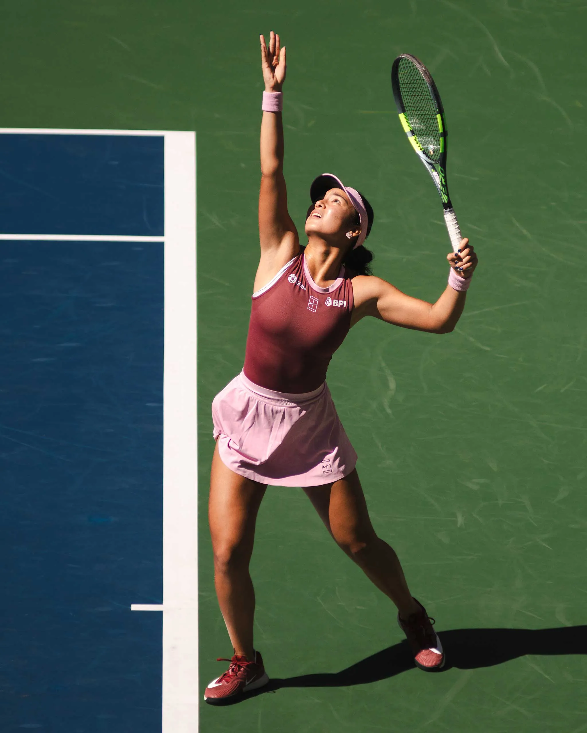 Female tennis player in pink outfit reaches upward with racquet in a high volley on a green court.