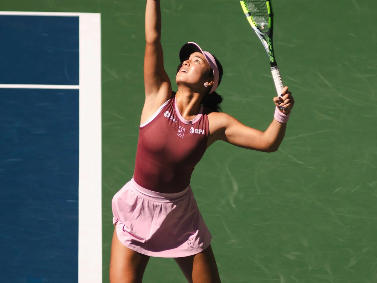 Female tennis player in pink outfit reaching up with her racket on a green court, mid-serve motion.