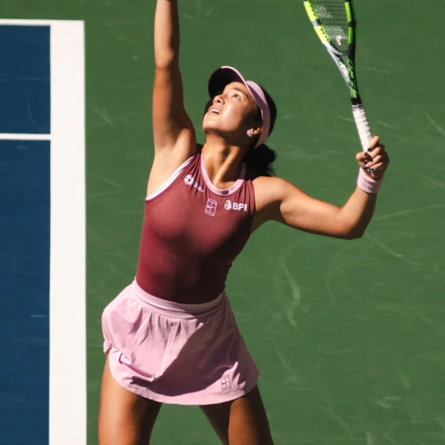Female tennis player in a pink outfit and visor reaching up with her racket on a green tennis court.
