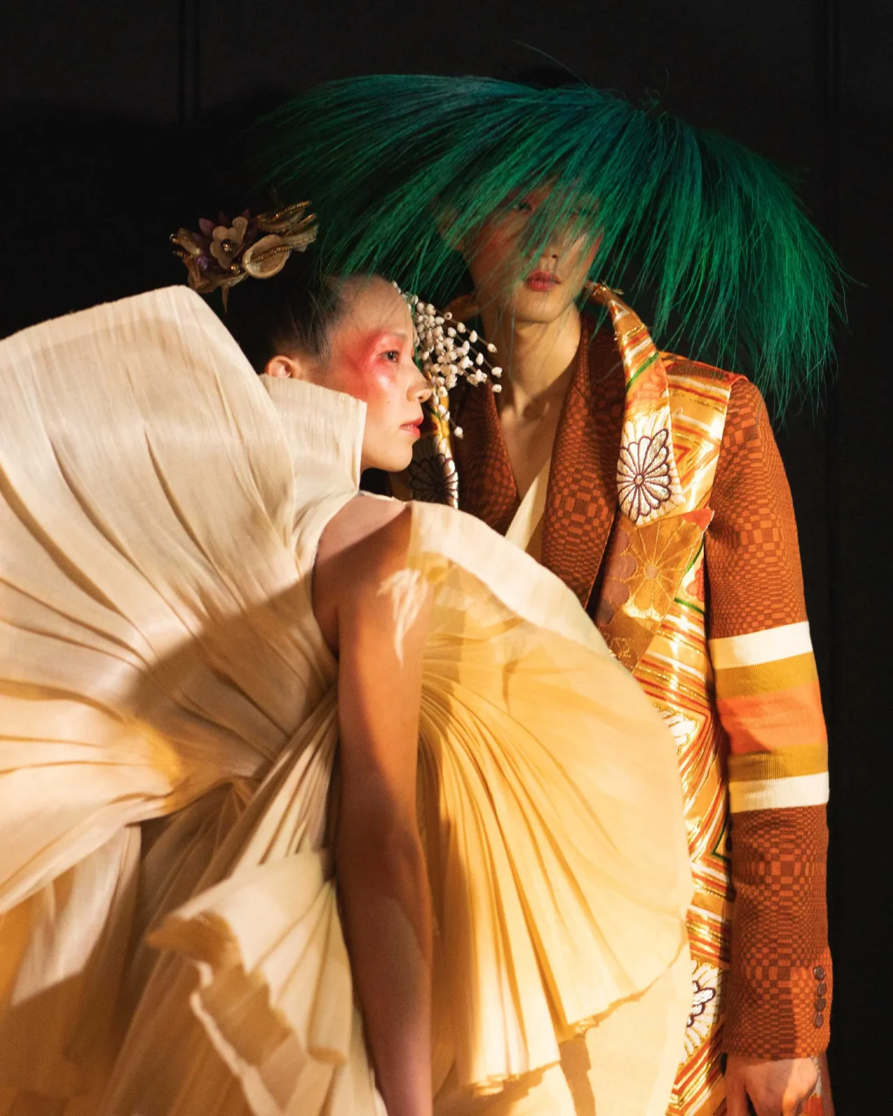 Line of models on a runway in bright, eclectic outfits with oversized neon wigs against a dark background.