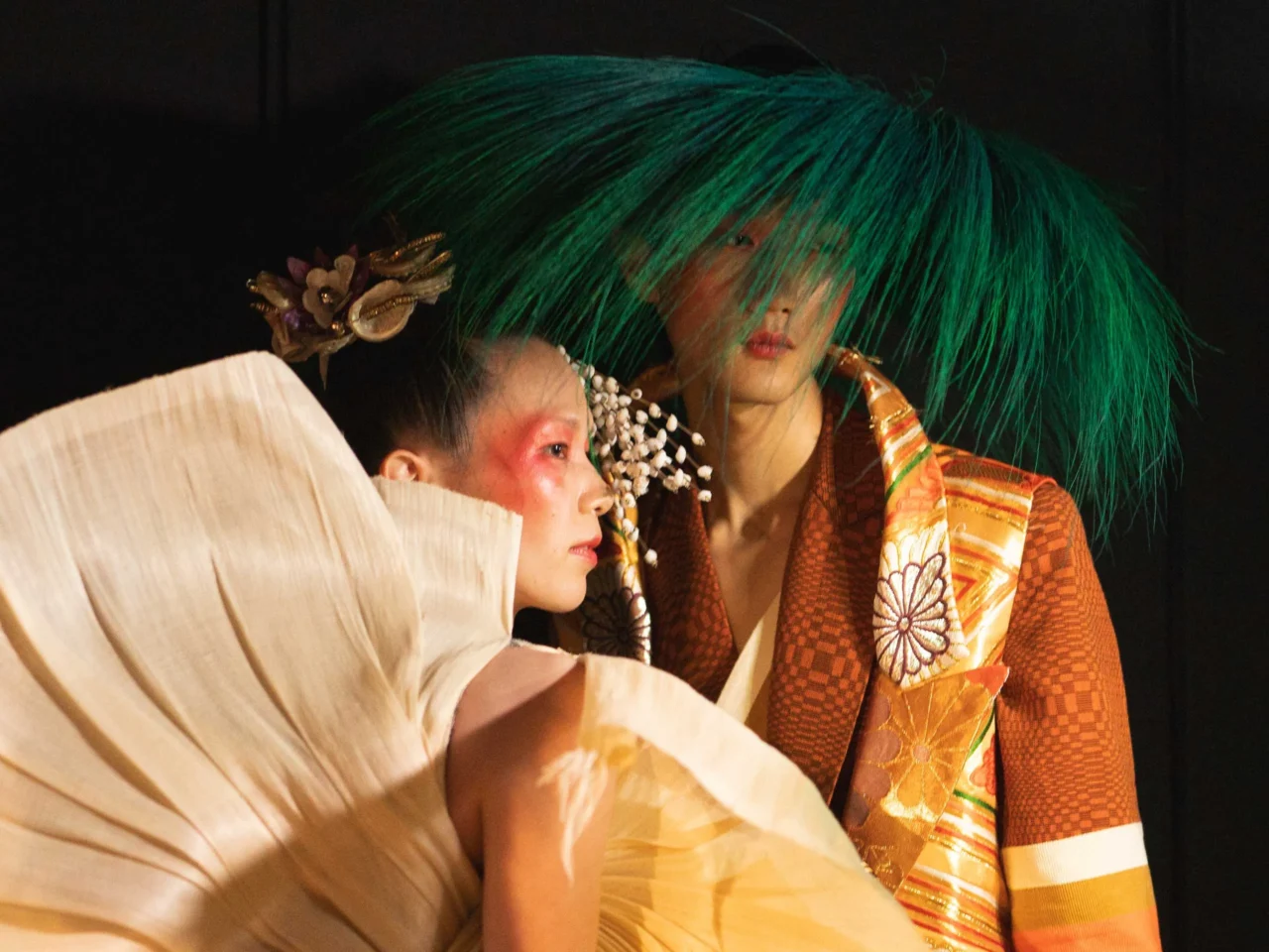 Line of models on a runway in bright, eclectic outfits with oversized neon wigs against a dark background.
