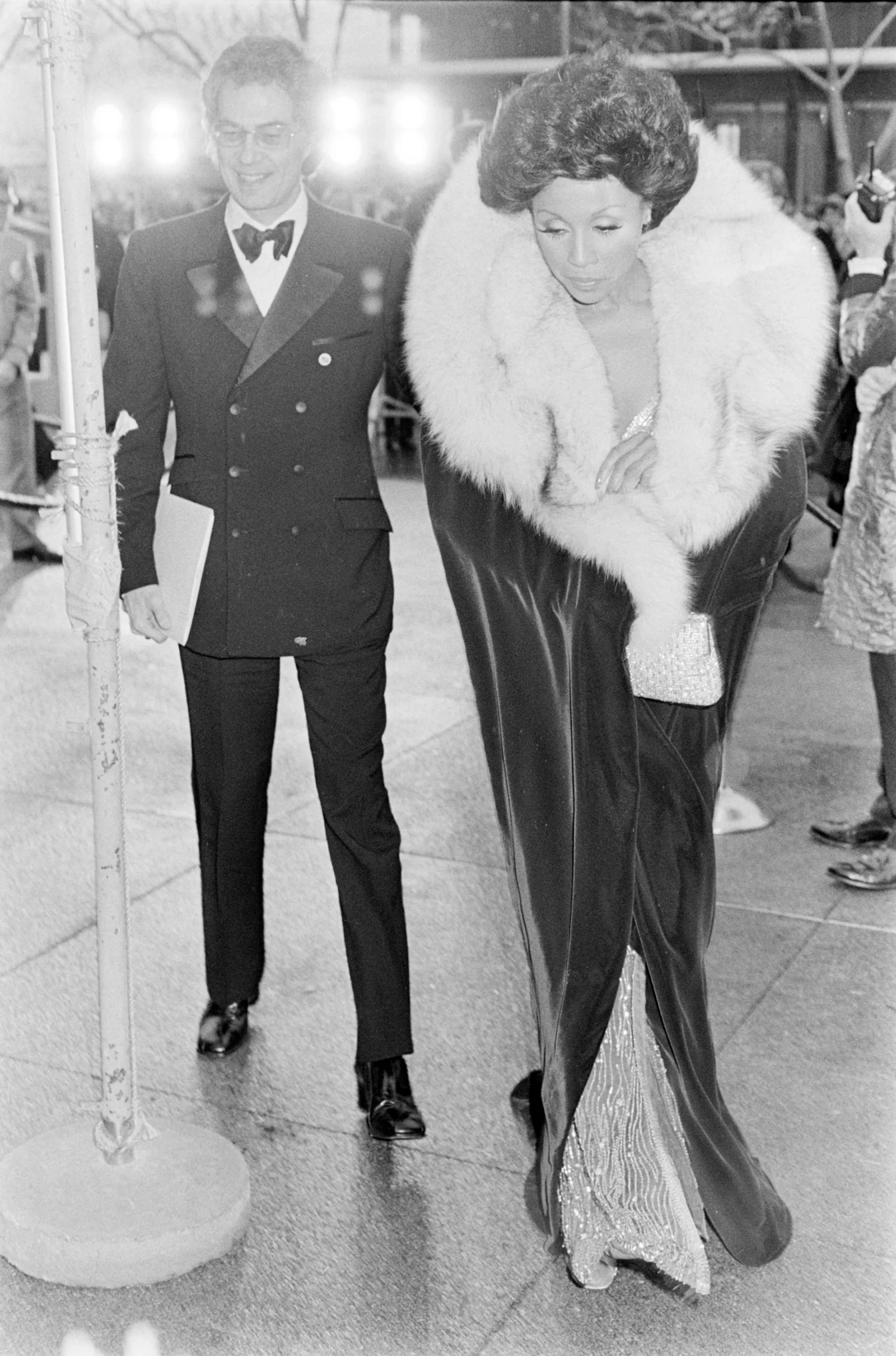 Man in a double-breasted tuxedo and a woman in a fur-trimmed gown walking arm in arm on a city sidewalk.