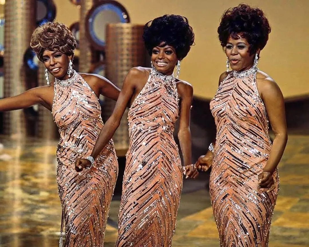 Three African American women in matching peach sequin gowns perform on a stage, mid-dance with smiles on their faces.