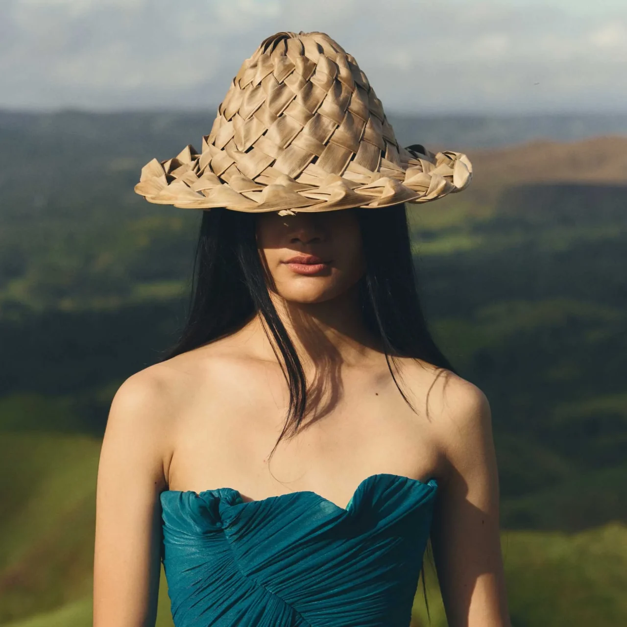 Woman in a teal dress standing on a sunlit grassy hill, with rolling green hills and distant misty mountains beyond you.