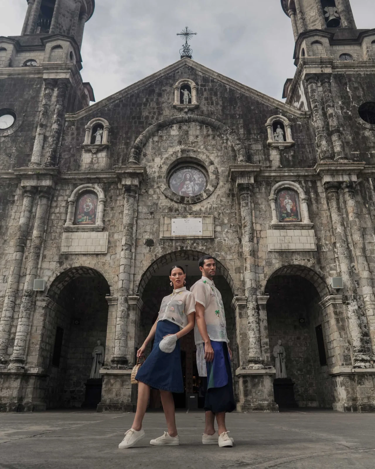 Two models stand back-to-back in front of a stone arched building, wearing light patterned shirts and blue pants, with one holding a woven hat.