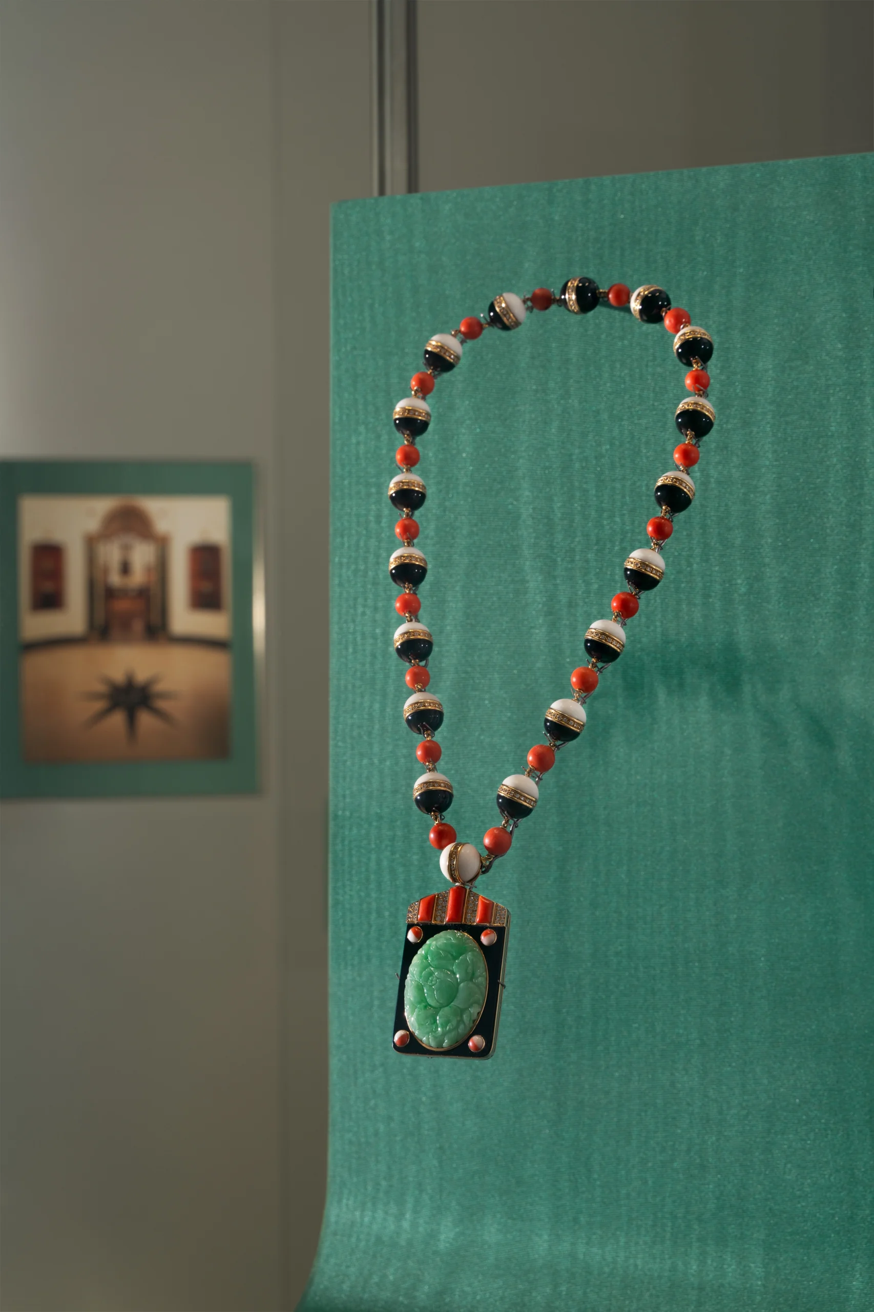Beaded necklace with red, white, and black beads and a green pendant on a teal display panel in a museum-esque setting.