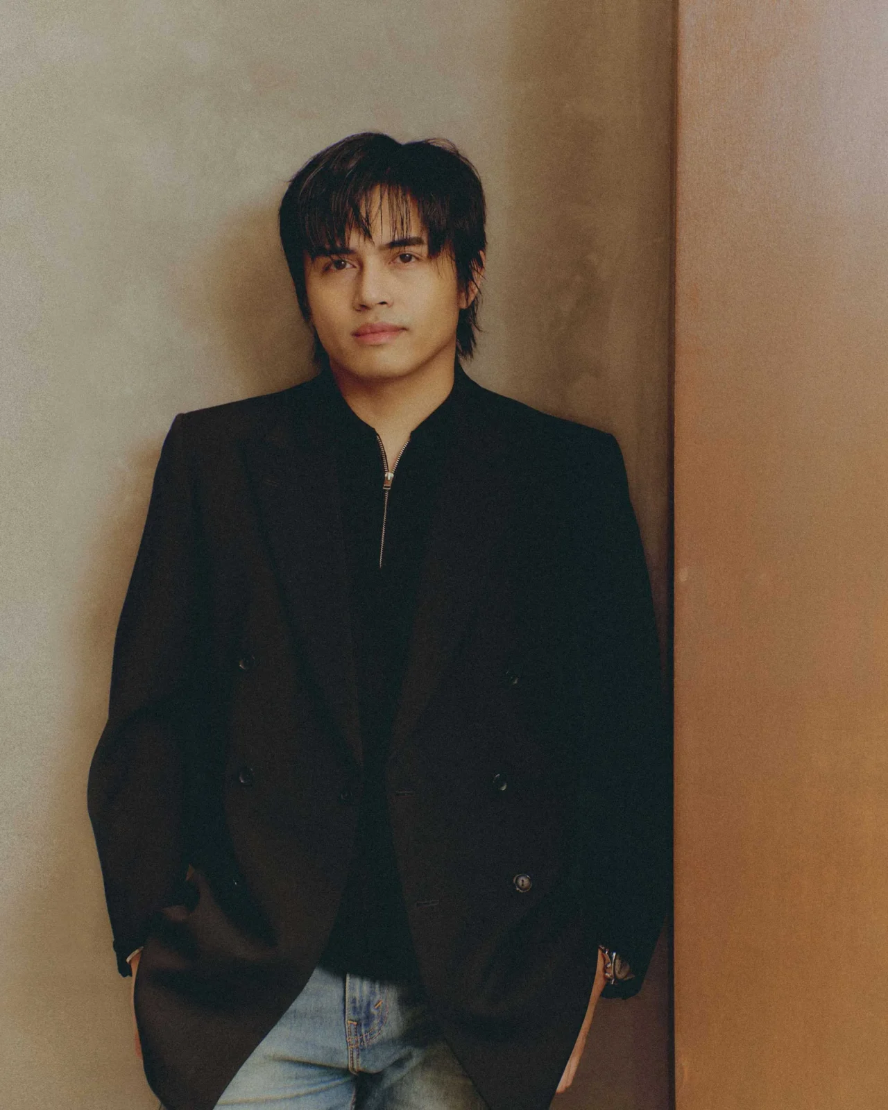 Portrait of a young man with dark, wavy hair wearing a black zip-up jacket, standing against a beige wall with a calm expression.